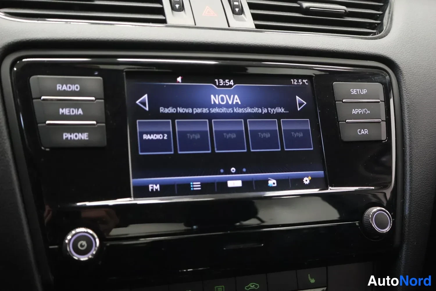 Yksityiskohta: Kojelaudan keskikäyttöpaneeli: kosketusnäyttö soittaa radioasemaa NOVA, vasemmalla RADIO-, MEDIA- ja PHONE-napit, oikealla SETUP-, APP- ja CAR-napit.