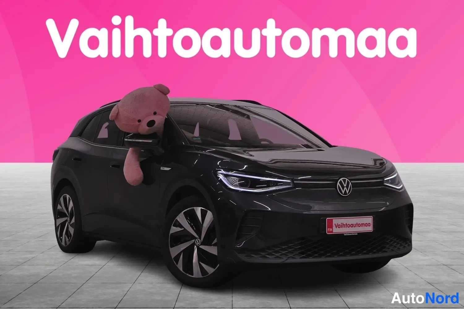 Etuviisto, nokka oikealle: Mustan Volkswagen-henkilöauton etunurkkikuva oikealta, keulasta ja kuljettajan puolen sivusta, taustalla vaaleanpunainen mainoslogo.