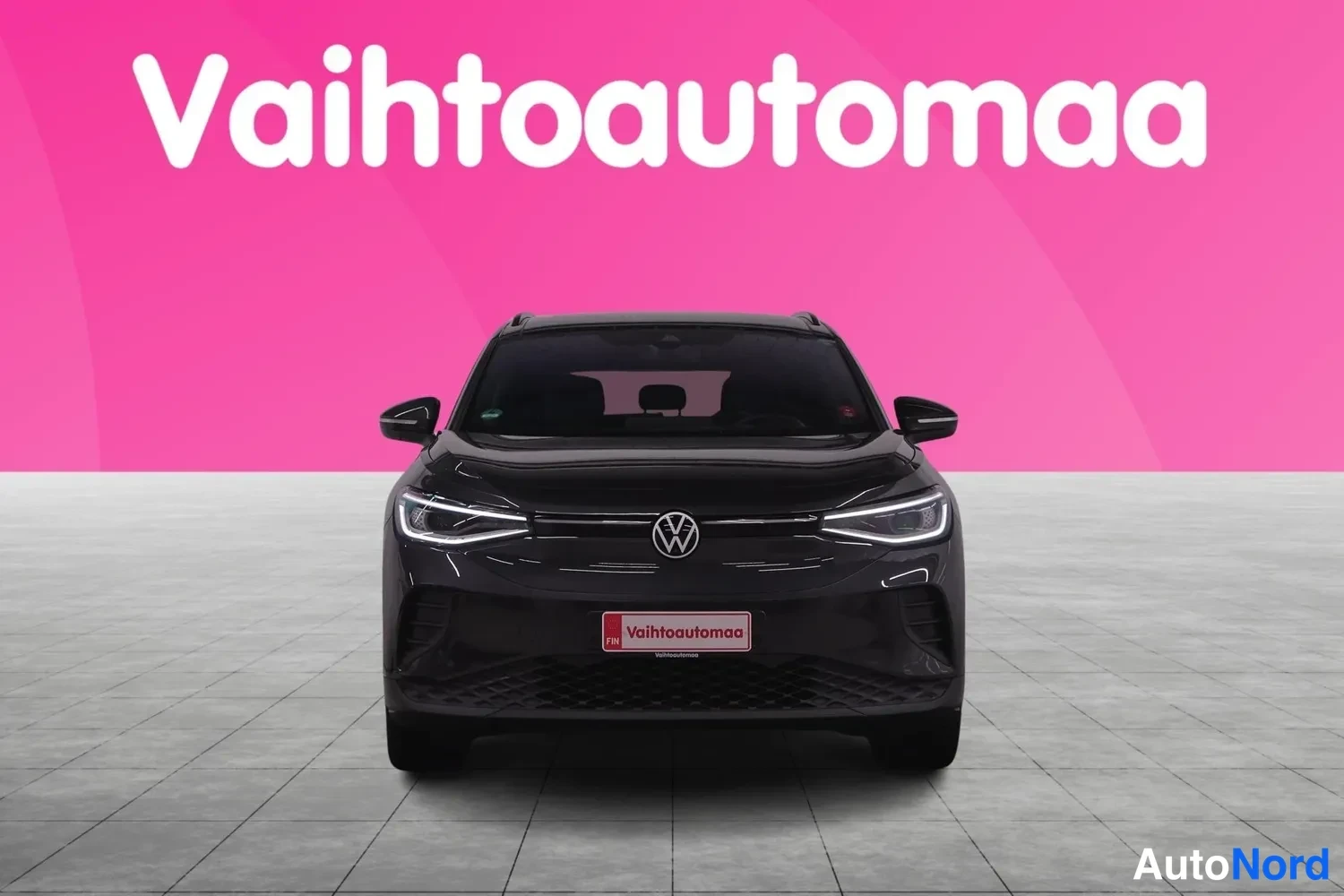 Edestä: Mustan Volkswagen-henkilöauton suora edessäpäinkuva, keulan etupansa, lyhdyt ja merkki näkyvät, taustalla vaaleanpunainen mainoslogo.