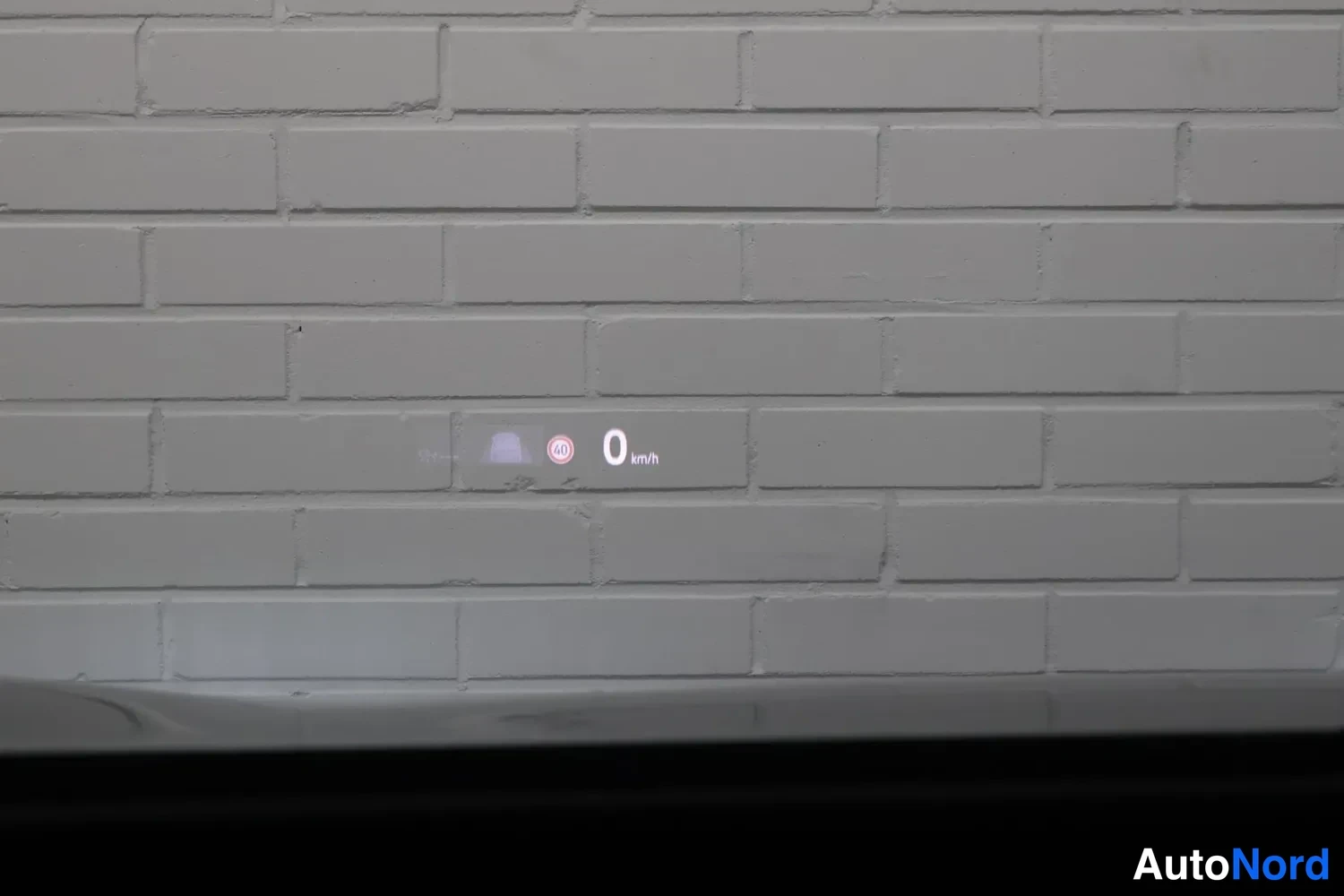 Muu: Tuulilasiprojektio (head-up display) heijastuu valkoisella tiiliseinällä: nopeus 0 km/h ja pyöreä 40-merkki.