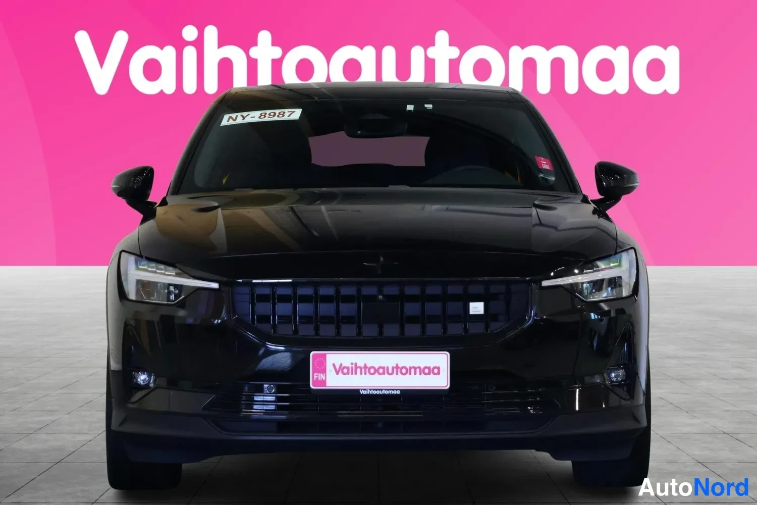 Edestä: Mustan Polestar 2:n suora etunäkymä, pinkki tausta ja Vaihtoautomaa-kyltti etupuskurissa.