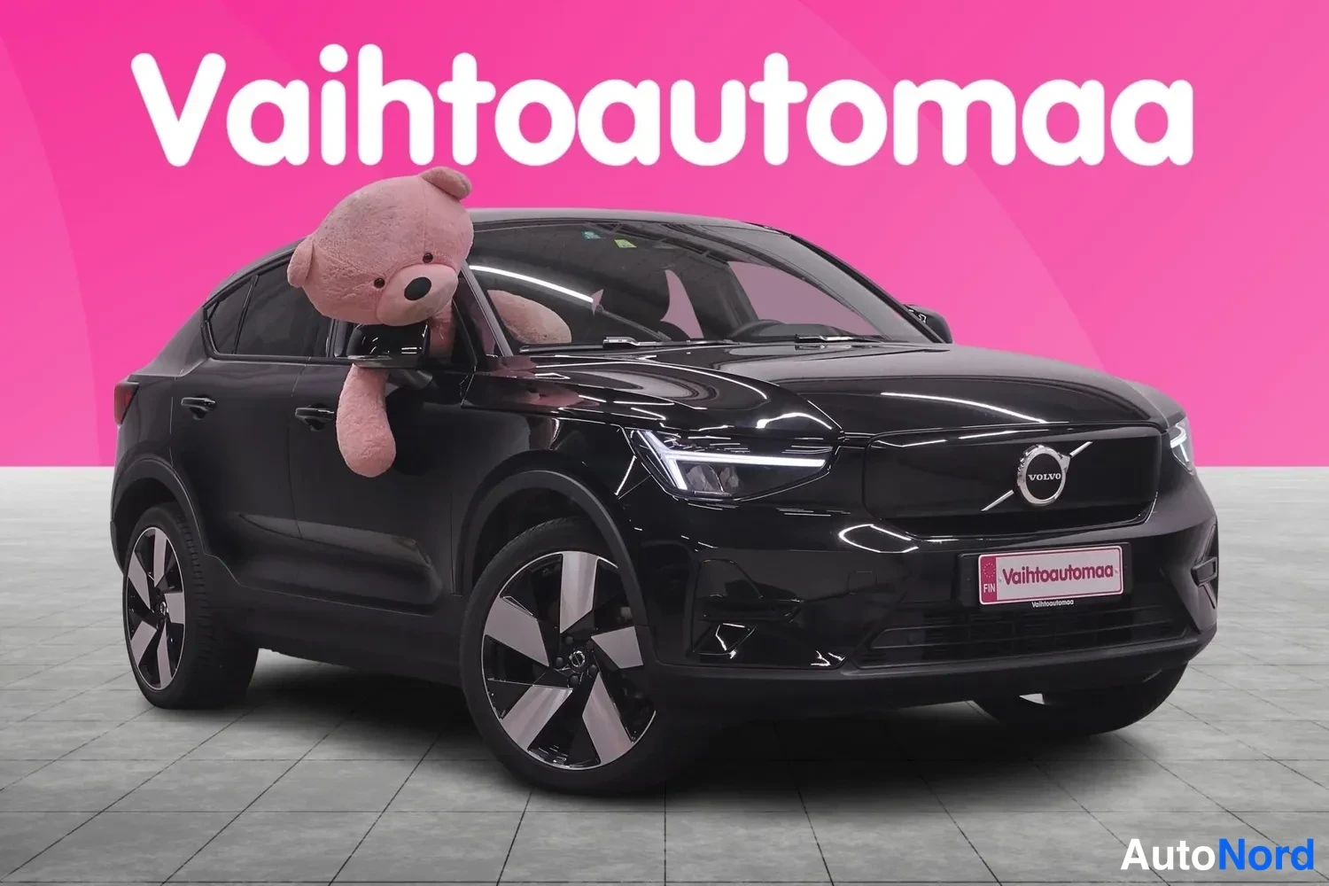 Etuviisto, nokka oikealle: Musta Volvo-henkilöauto etu-kulmasta oikealta nähden, keulakoriste ja etupyörät näkyvät, suuri pehmolelu takanäytössä.