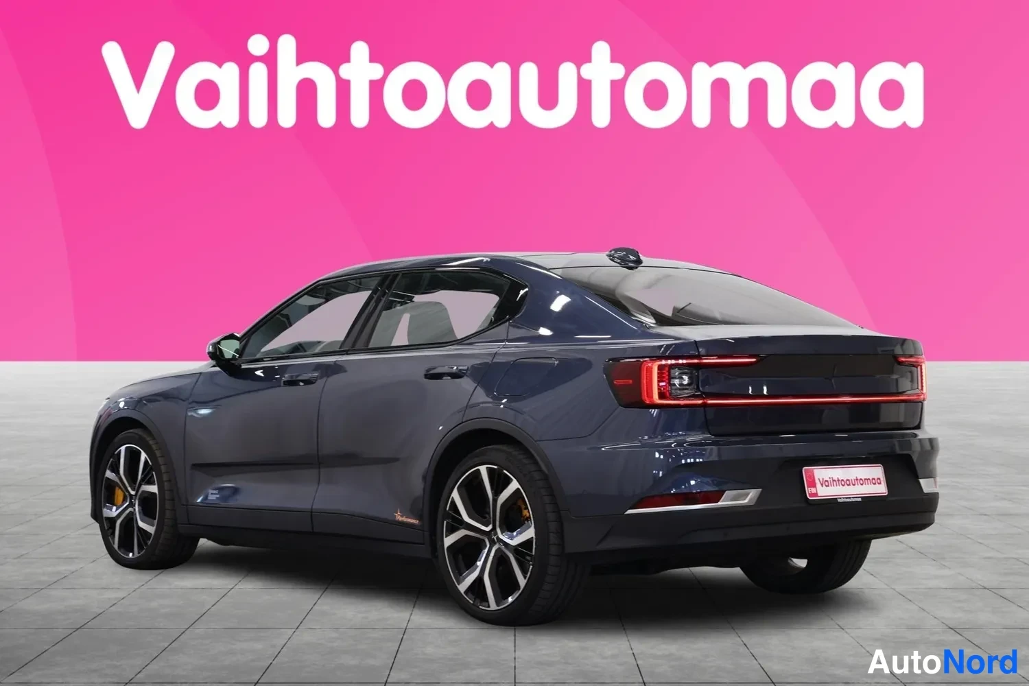 Takaviisto: Tummansininen Polestar 2 takaviistosta vasemmalta kuvattuna studiossa; näkyvät kattospoileri, poikittainen takavalo ja kevytmetallivanteet.