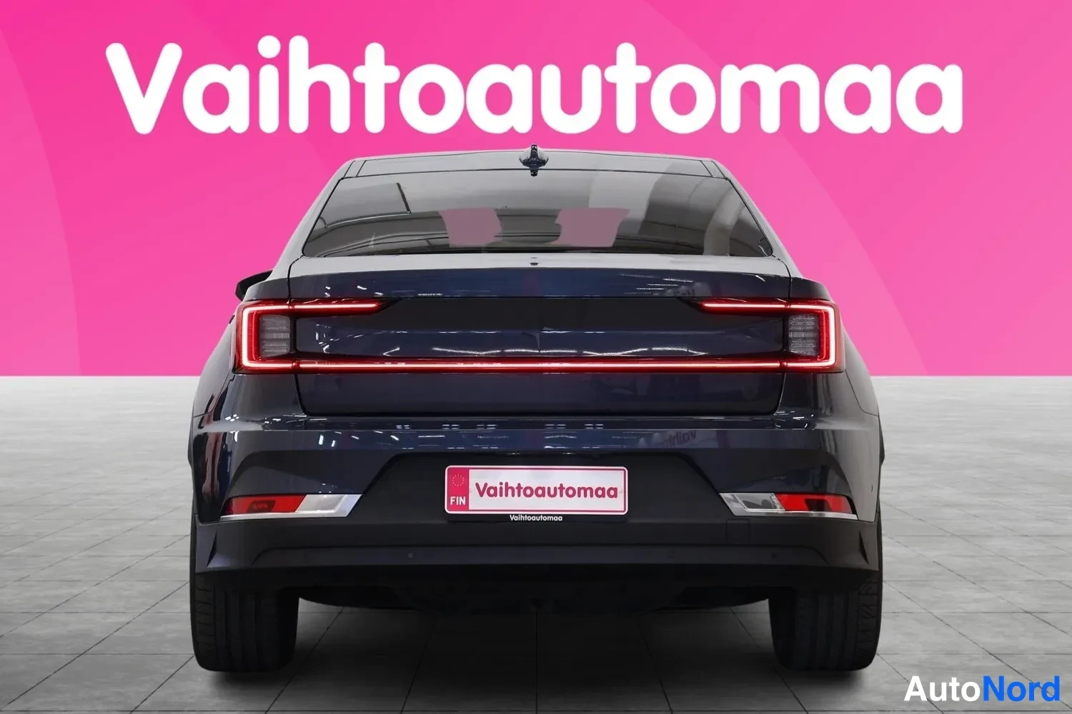 Takaa: Sinertävän tumman sedanin takaosan suora etänä, koko leveydeltään avautuva punainen takavalojennauhe ja Vahtoautomaa-kyltti rekisterin paikalla.