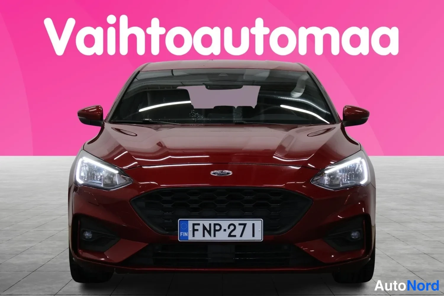 Edestä: Punaisenruskean Ford Focus -henkilöauton etupää suoraan edestä kuvattuna, rekisteritunnus, LED-päiväajovalot päällä.