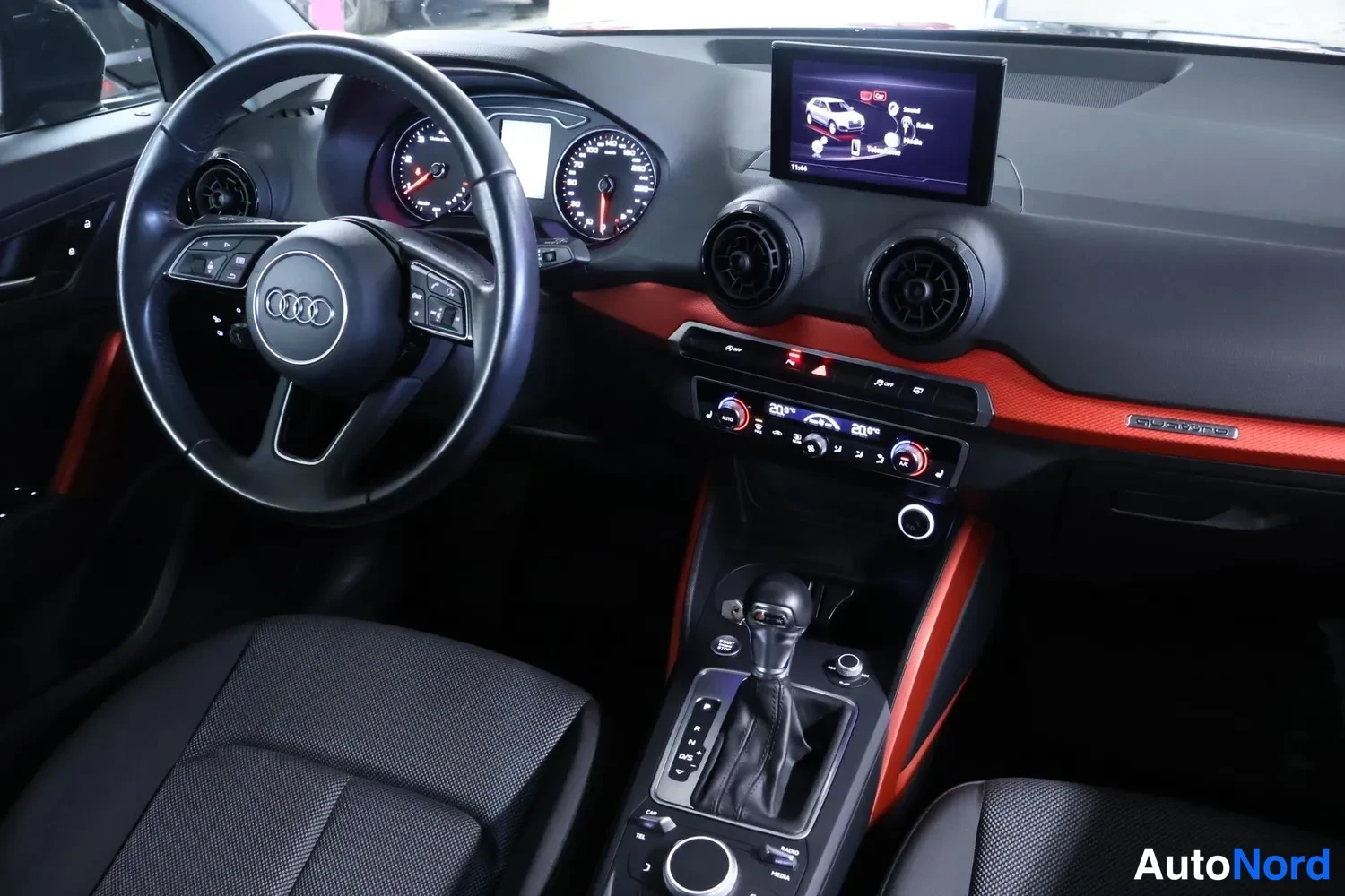 Etuviisto, nokka vasemmalle: Audi Q2:n ohjaamo kuljettajan puolelta: nappiratti, ympyräilmastointisuihkut, punaiset koristelistat, automaattivaihteiston valitsin ja kosketusnäyttö.