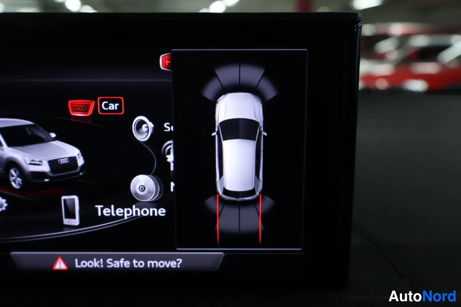 Yksityiskohta: Infotainment-näyttö näyttää pysäköintitutkan ylhäältäkuvan autosta punaisilla ohjausviivoilla; näkyvissä myös valikot 'Car' ja 'Telephone' sekä varoitusteksti '