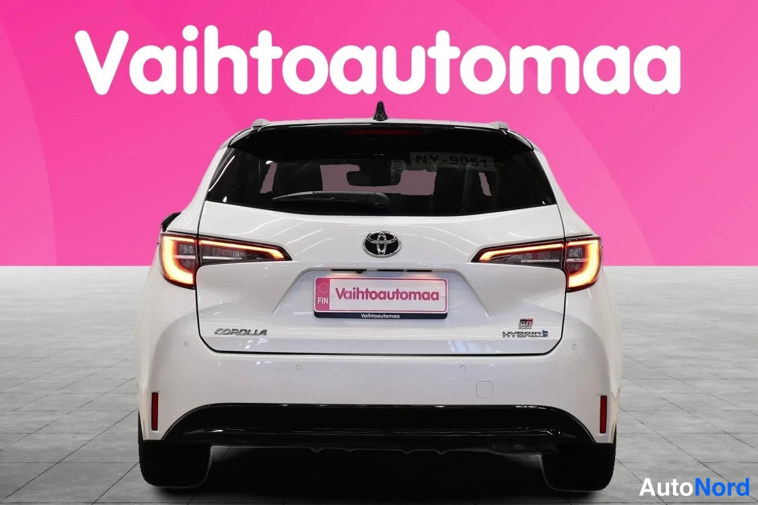 Takaa: Valkoisen farmariauton takaosa suoraan takaa: Toyota-merkki, COROLLA-teksti, Hybrid-tunnus ja punaiset takavalot.