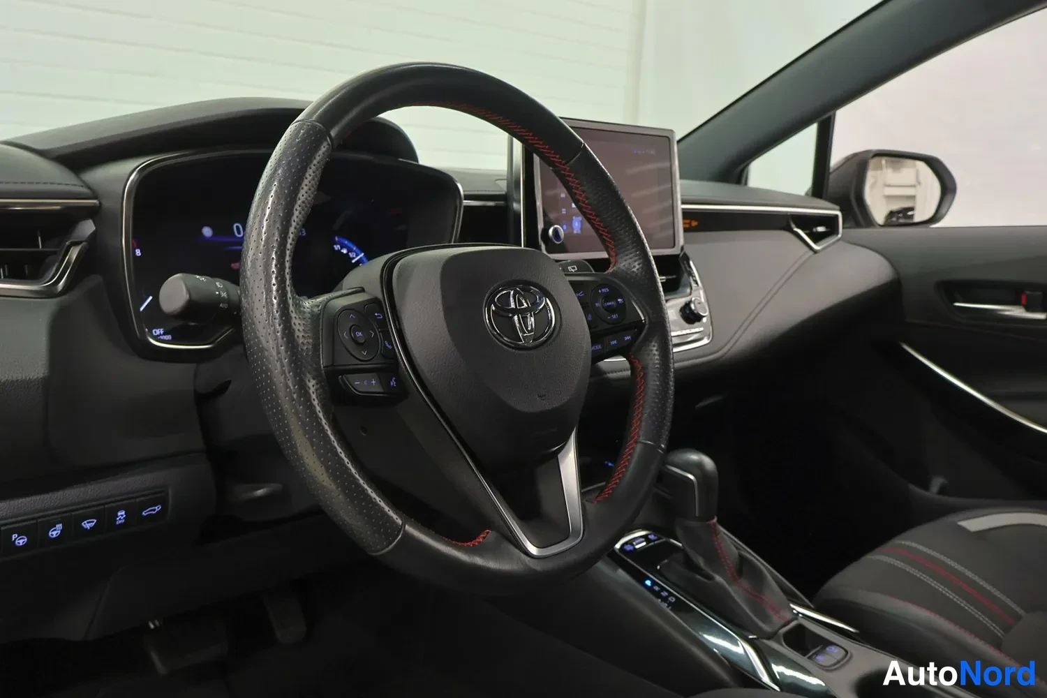Edestä: Ohjauspyörä Toyota-logolla ja punaisilla ompeleilla, mittaristo ja kosketusnäyttö keskellä.