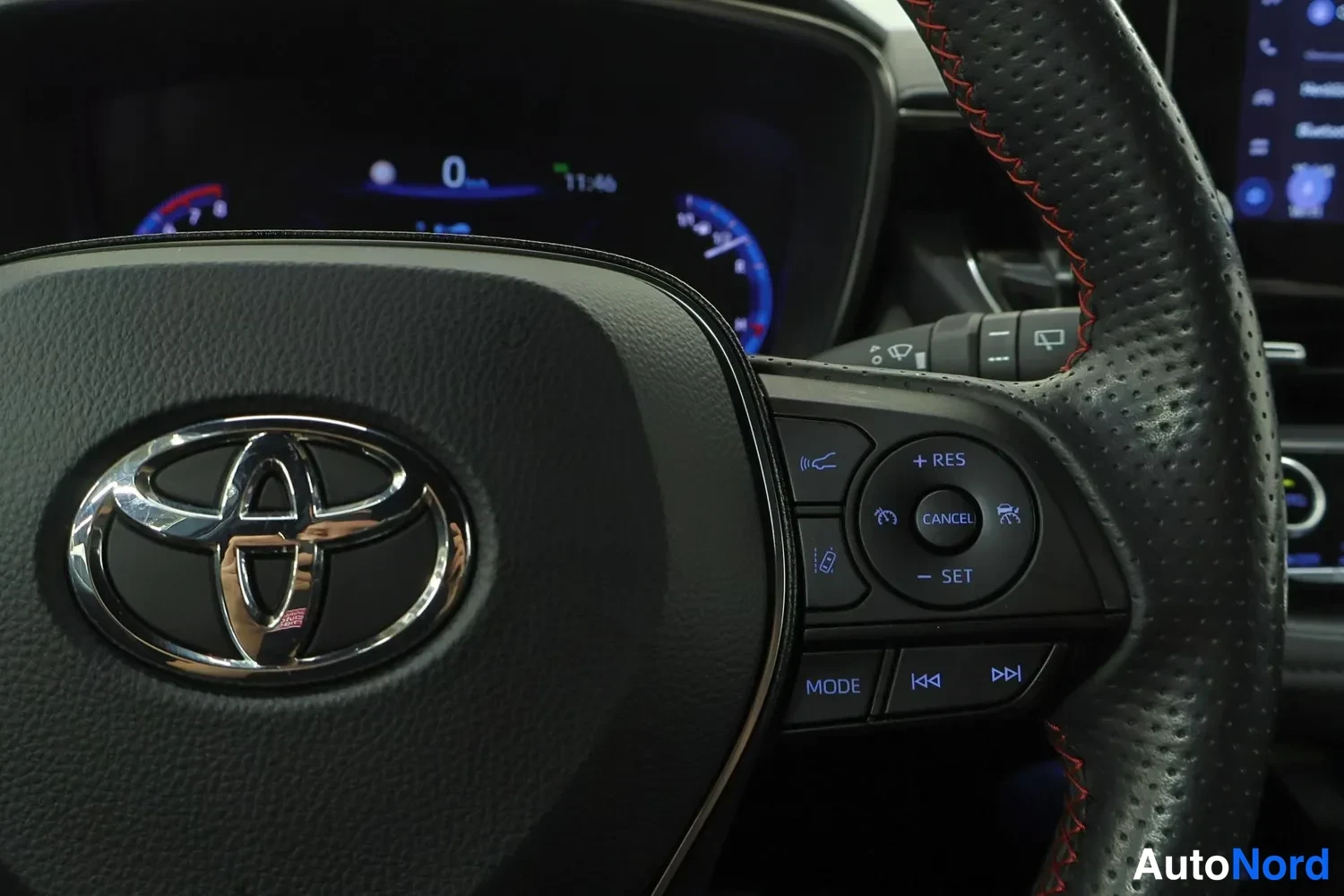 Yksityiskohta: Ohjauspyörän nærkuva Toyota-merkillä ja vaa'alla ommelluilla punaisilla ompeleilla; oikealla puolella pikanäppäimet MODE ja mediatoiminnot.