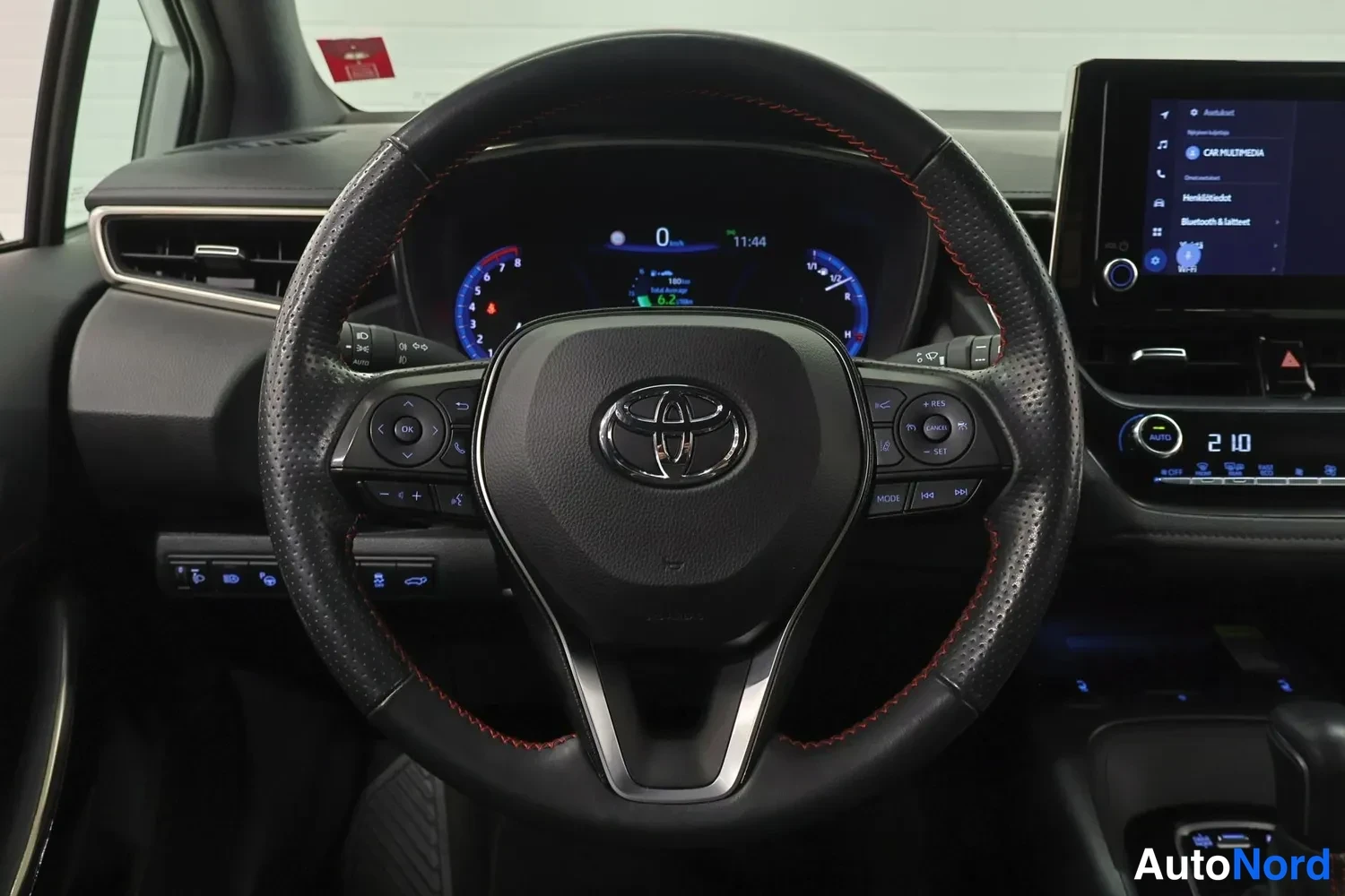 Yksityiskohta: Ohjaustehostimen taustalla kojelaudan mittarit, keskellä Toyota-merkki ja punaiset ompeleet; oikealla kosketusnäyttö ja lämpötilanäyttö 21,0.