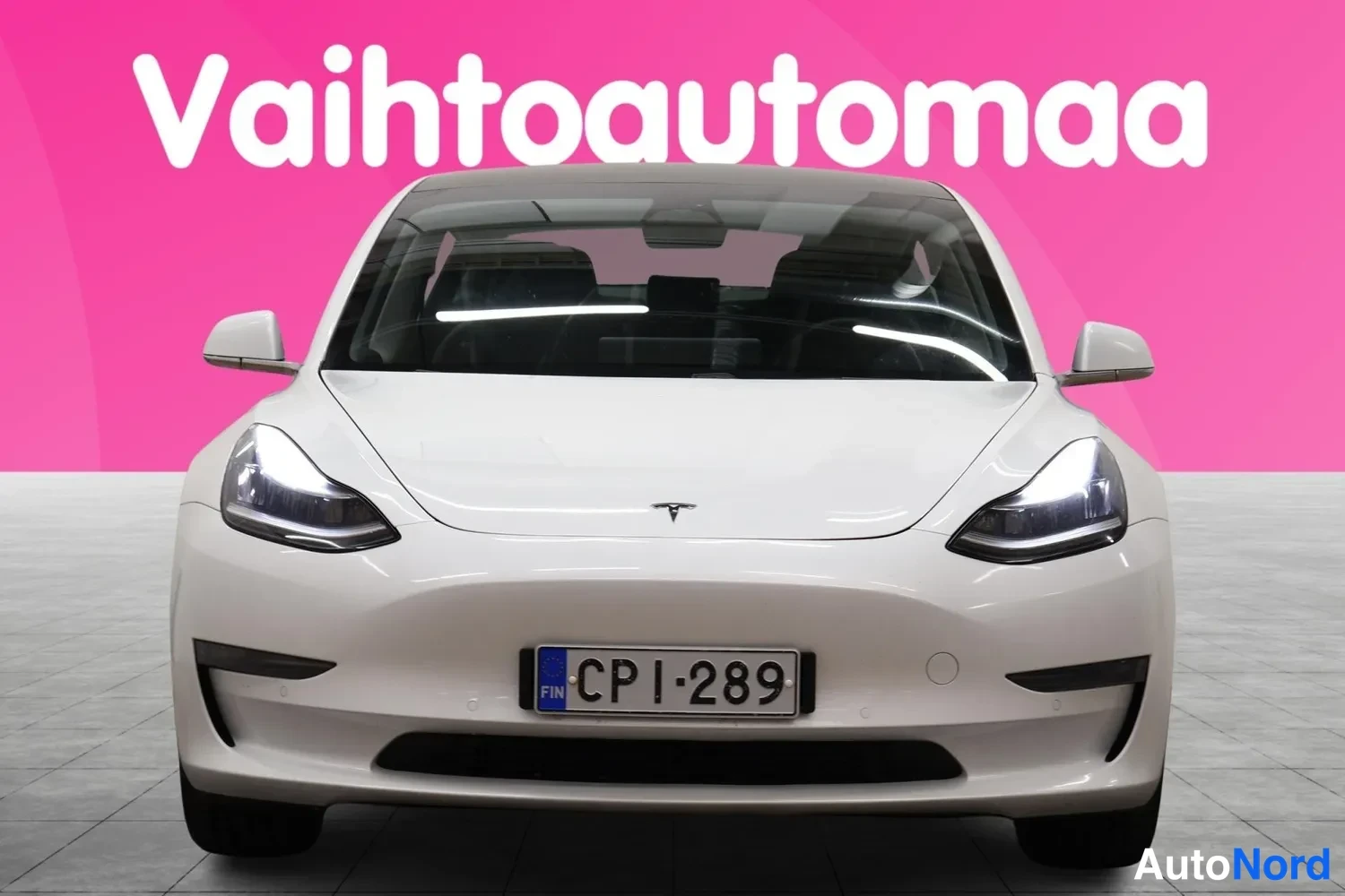 Edestä: Straight-on frontal view of a white Tesla Model 3 in a studio with a pink background. The LED headlights and license plate are centered.
