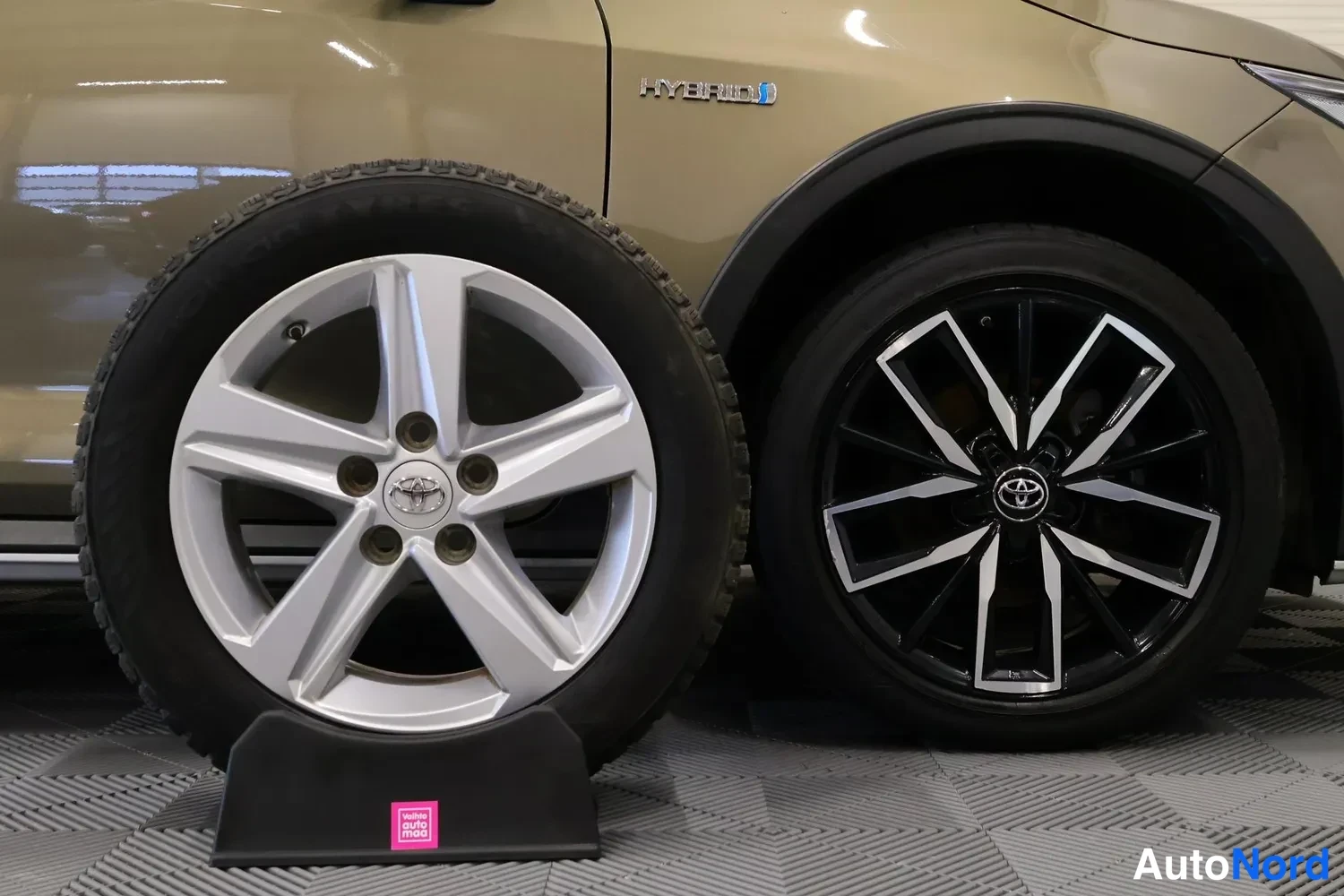 Yksityiskohta: Close-up of the front-left side of a beige car showing one spare tire with silver alloy wheel leaning against the vehicle and another wheel mounted on the car;