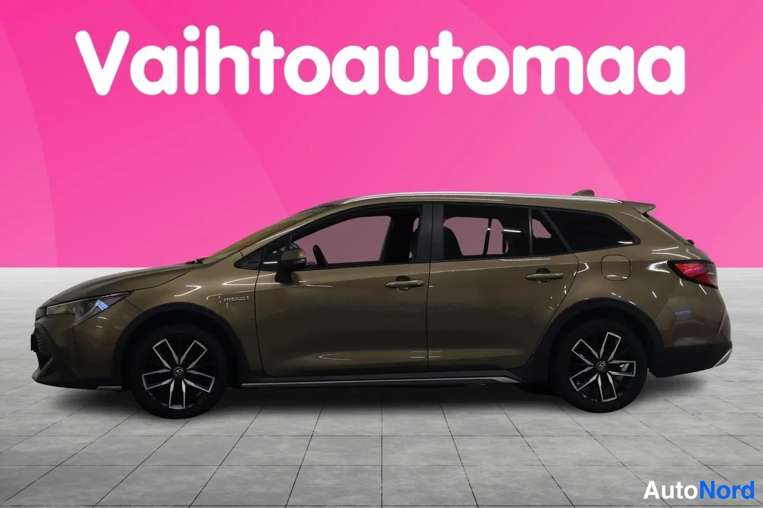 Sivulta: Toyota Corolla Touring Sports -farmariauto sivulta vasemmasta suunnasta, ruskean-kultainen koriväri, mustat kevytmetallivanteet, vaalea harmaa studio ja pinkki