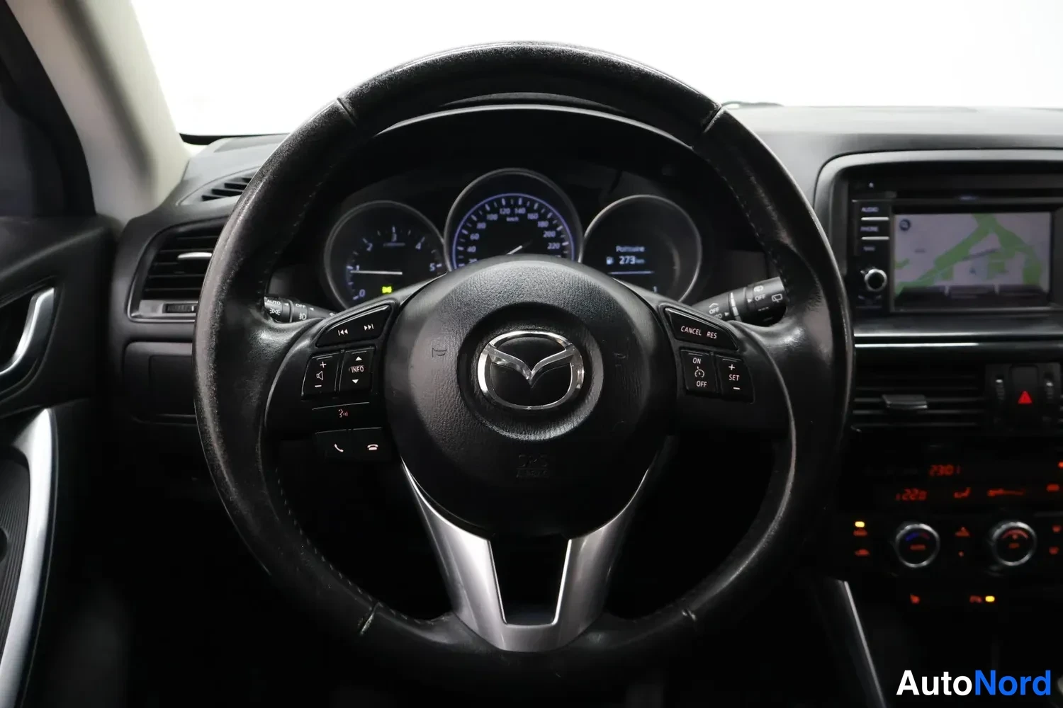Edestä: Ohjauspyörä ja mittaristo edestä kuvattuna, keskusnäytössä kartta, Mazda-tunnuksella.