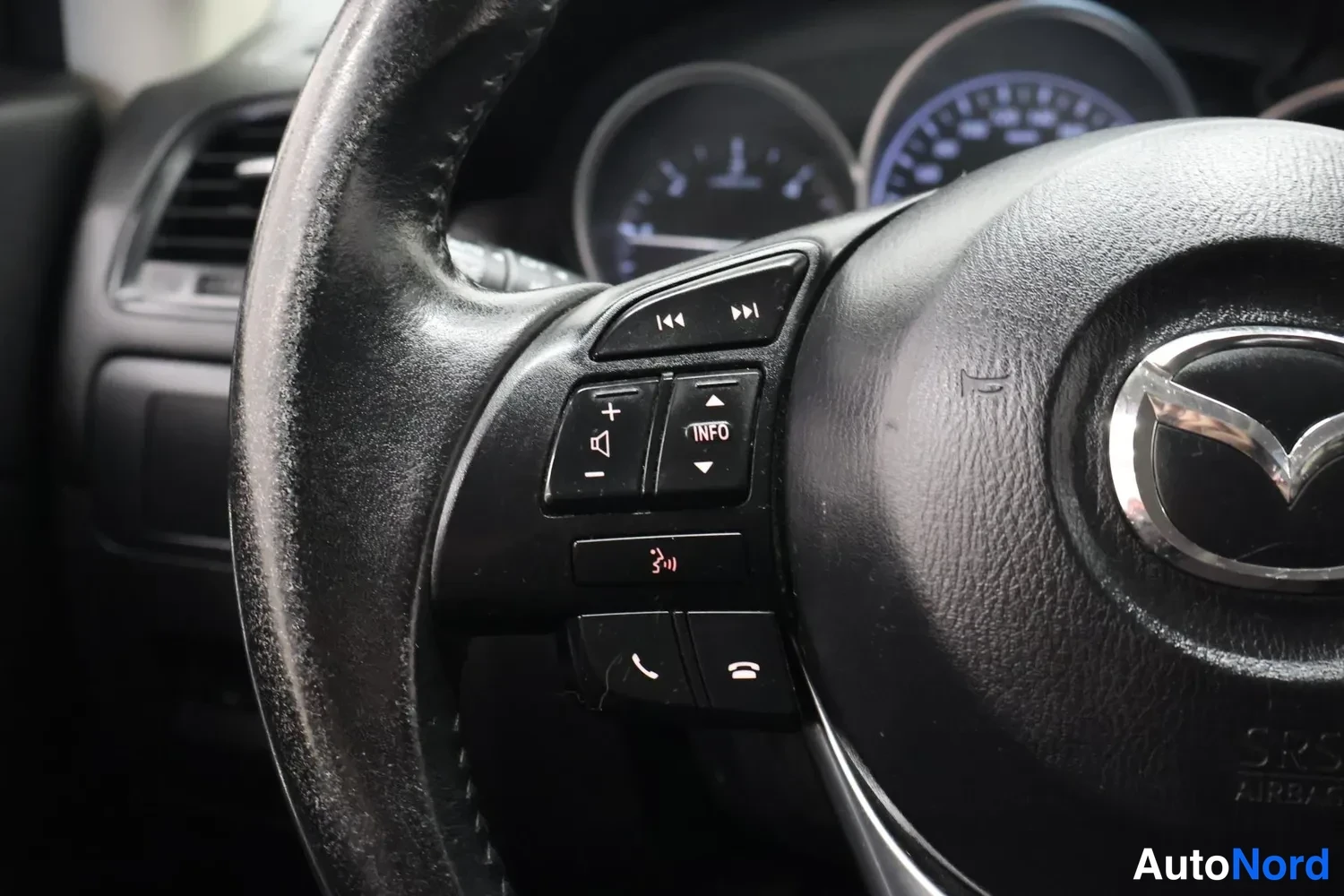 Muu: Lähikuva Mazda-ohjauspyörän vasemmasta puolesta: äänentoiston ja puhelun säätöpainikkeet, taustalla mittariston taulut.
