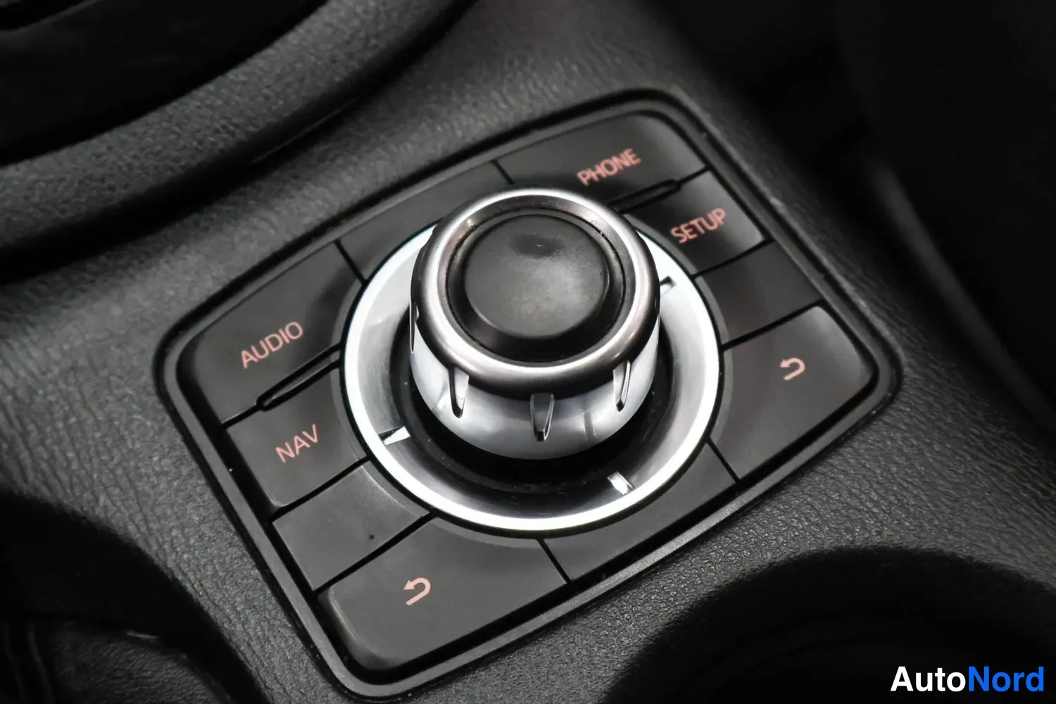 Yksityiskohta: Sisätilojen lähikuva auton pyöreästä infotainment-ohjausnupista, jonka ympärillä ovat PHONE-, SETUP-, AUDIO- ja NAV-painikkeet sekä paluunuoli-symbolit.