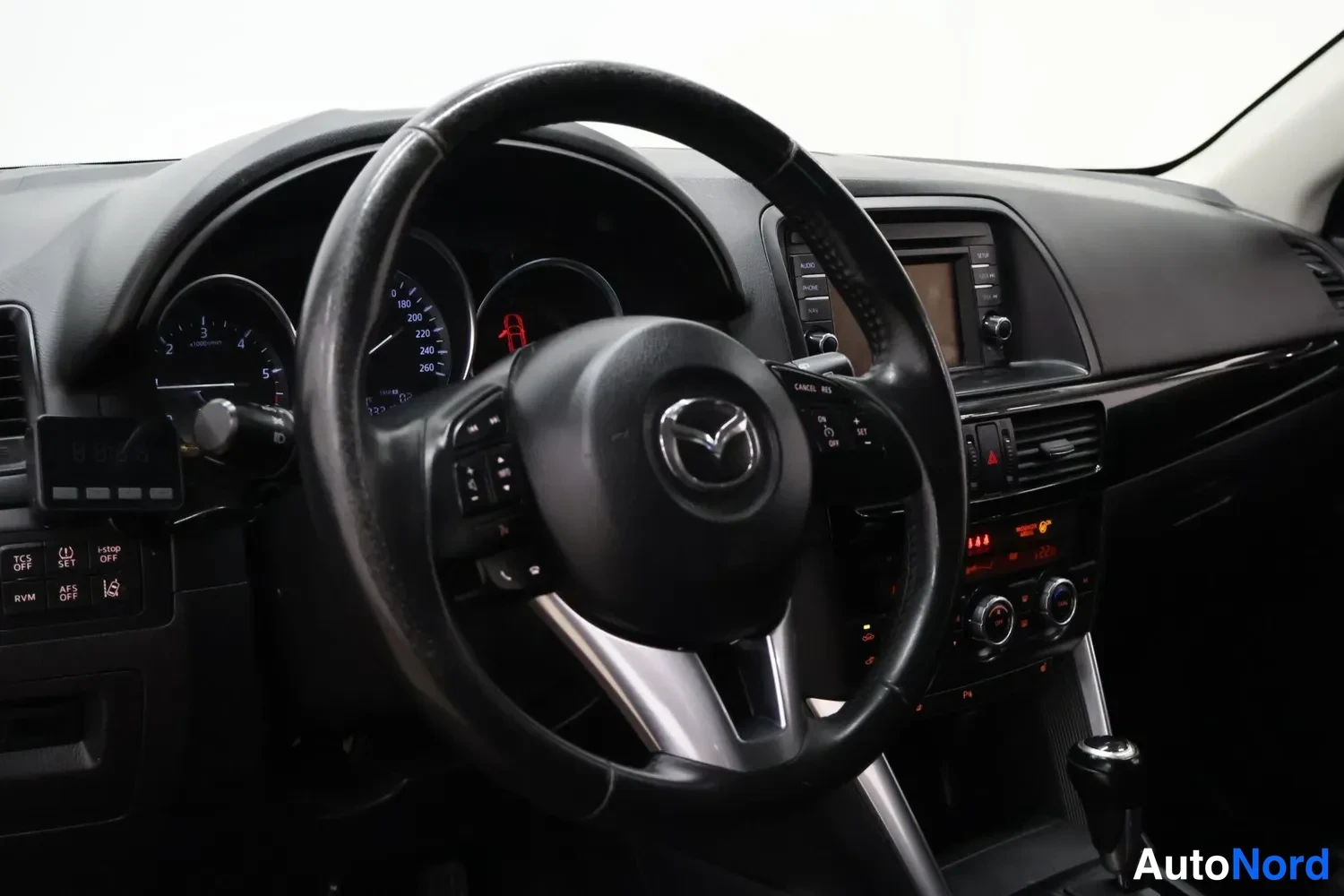 Edestä: Kuljettajan näkökulma Mazda-ohjauspyörään, mittaristo ja keskusnäyttö näkyvät.