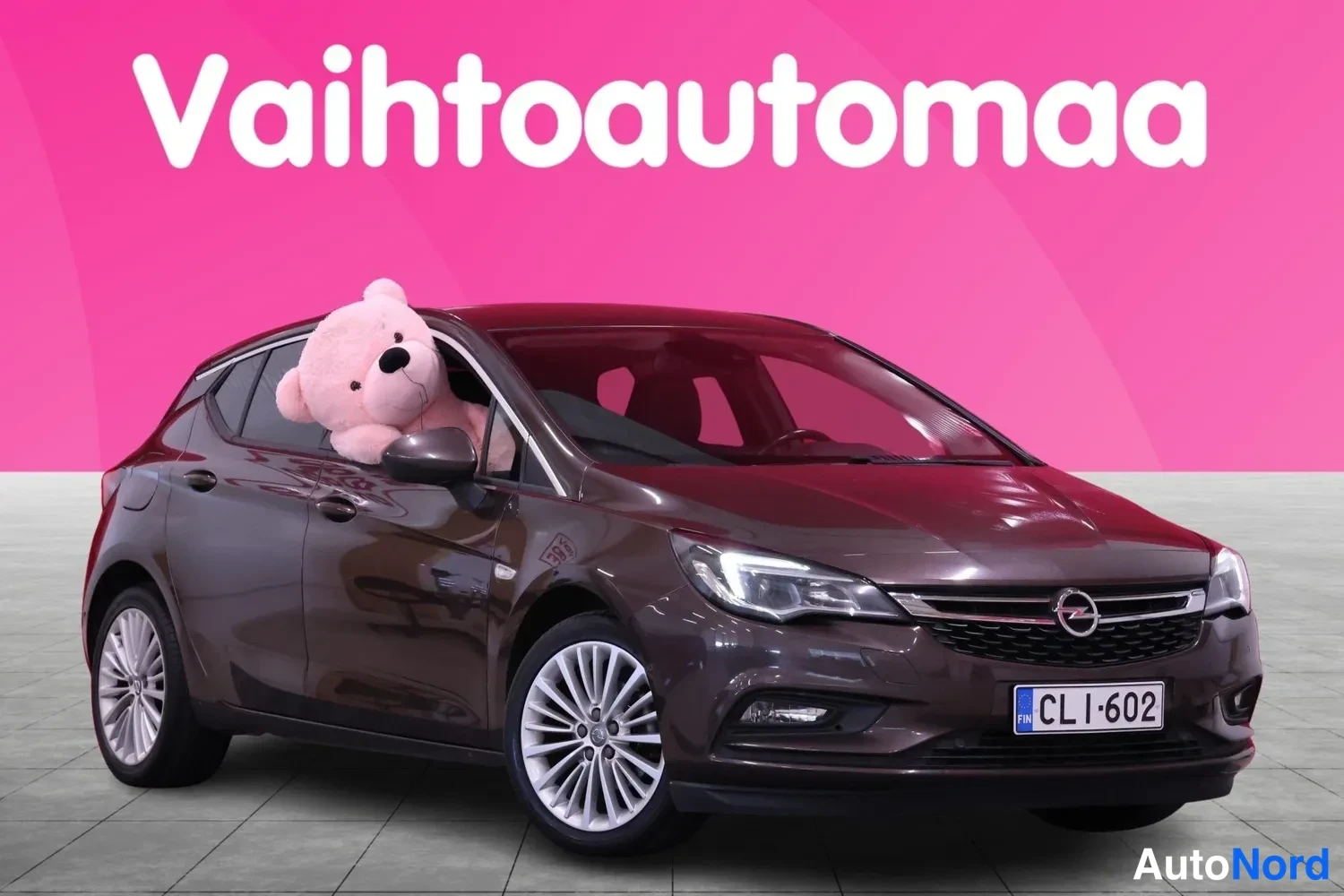 Etuviisto, nokka oikealle: Tummanruskea Opel Astra etuviistosta oikealta, etusivu vaaleilla vanteilla rekisterillä CL I-602, taustalla pinkki Vaihtoautomaa-banneri.