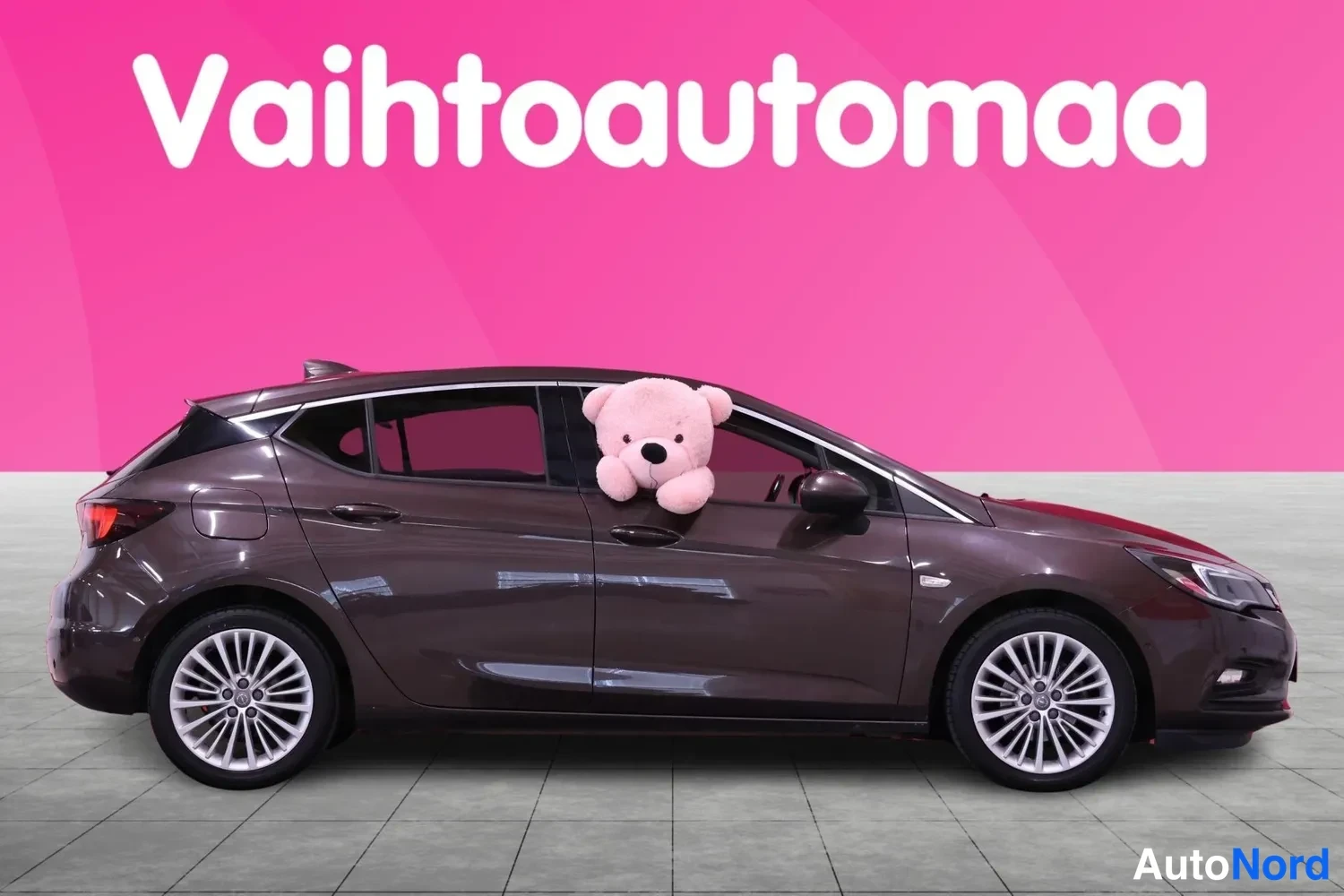 Sivulta: Tummanruskea Opel Astra sivulta oikealta, vaaleat vanteet ja takana spoileri, taustalla pinkki Vaihtoautomaa-banneri.