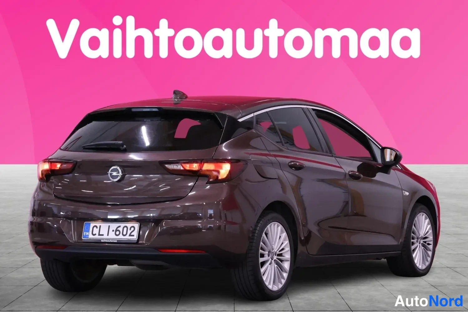 Takaviisto: Tummanruskea Opel Astra takaviistosta vasemmalta, takavalot ja rekisteri CL I-602 näkyvissä, taustalla pinkki banneri.