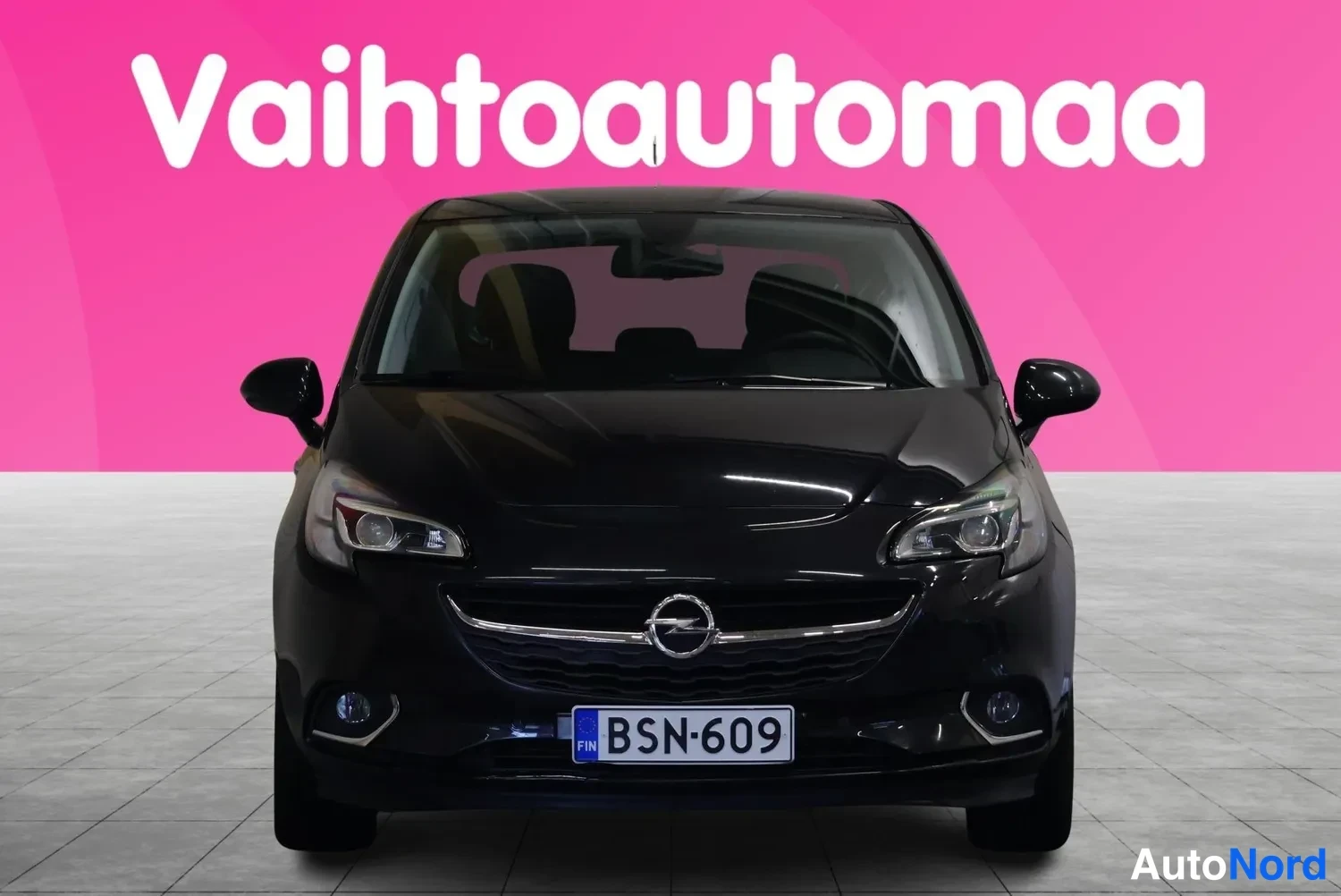 Edestä: Mustan hatchbackin suora etunäkymä, rekisterikilpi, pinkki tausta.