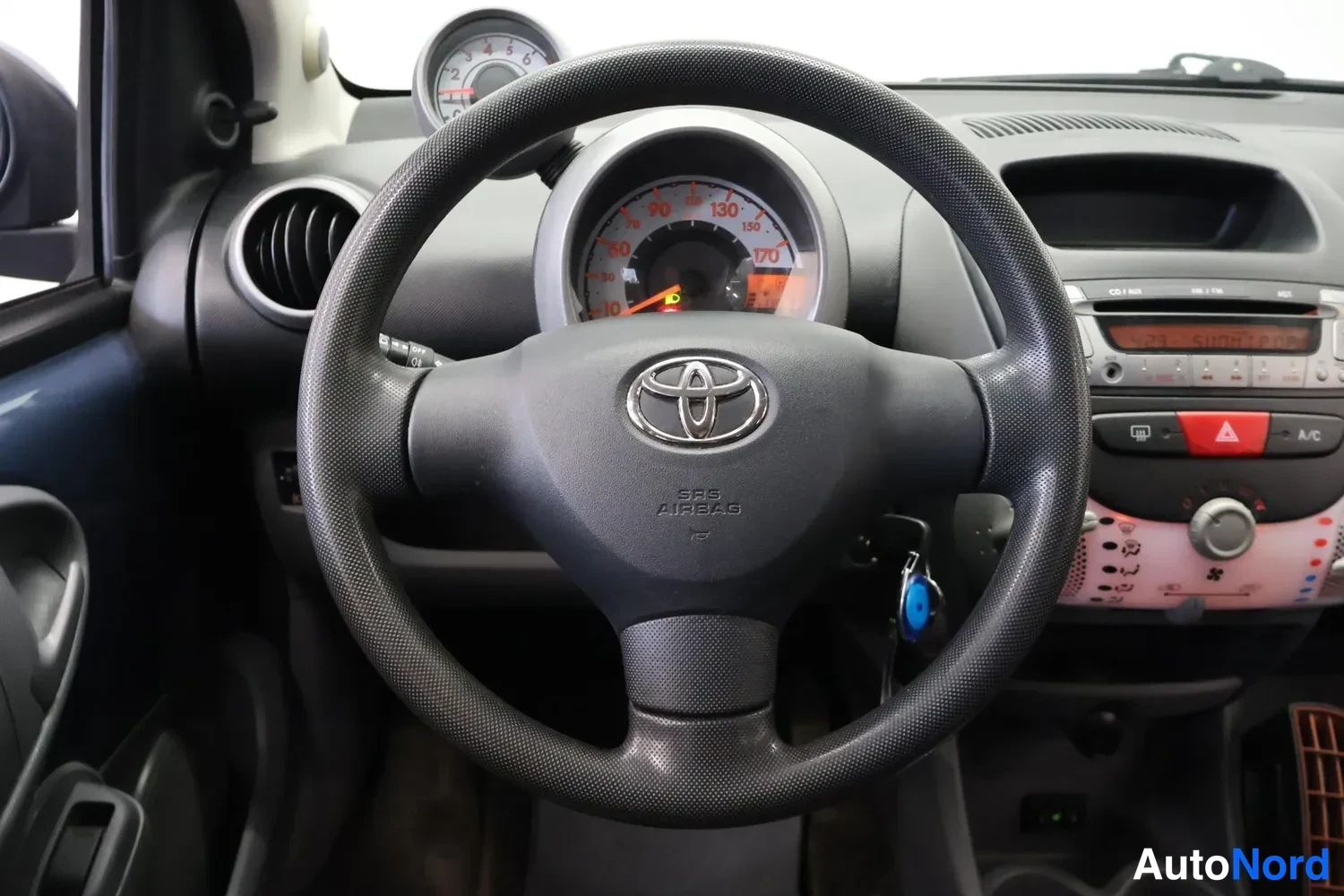 Muu: Ohjauspyörän lähikuva Toyotan merkillä ja SRS AIRBAG -tekstillä; taustalla mittaristo, ilmastointipaneeli, sininen avain lukossa ja A/C-painike näkyvissä.
