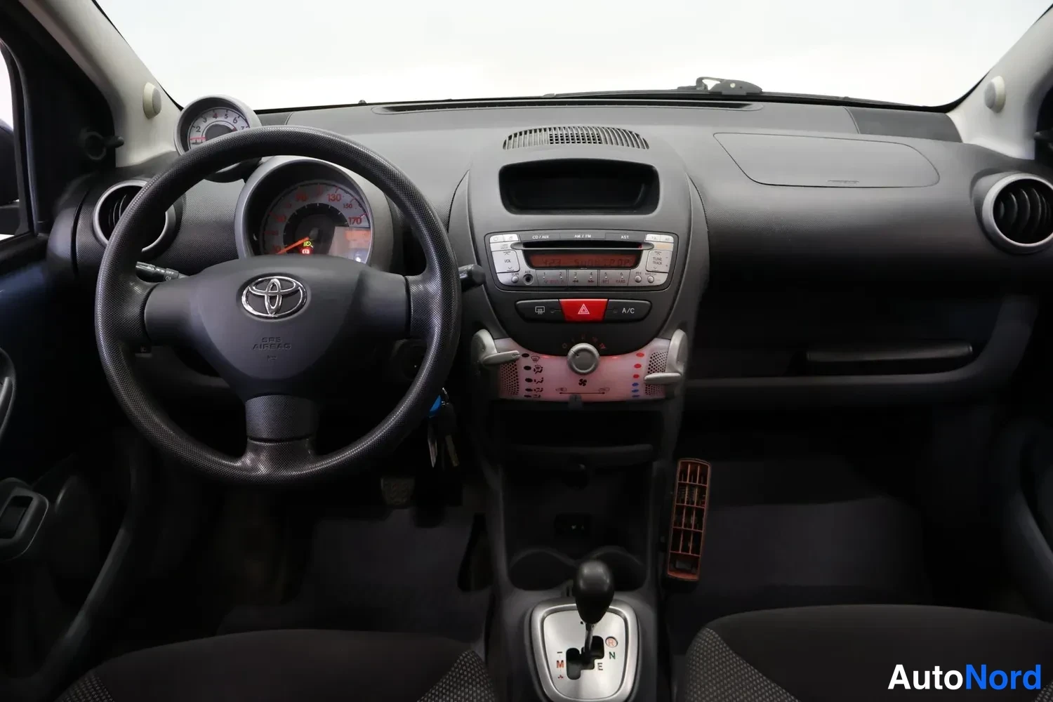 Muu: Kojelauta suoraan edestä: Toyota-ohjauspyörä ja mittarit vasemmalla, radio- ja ilmastointipaneeli keskellä, automaattivaihteiston valitsin M-, N-, R- ja E-asemi