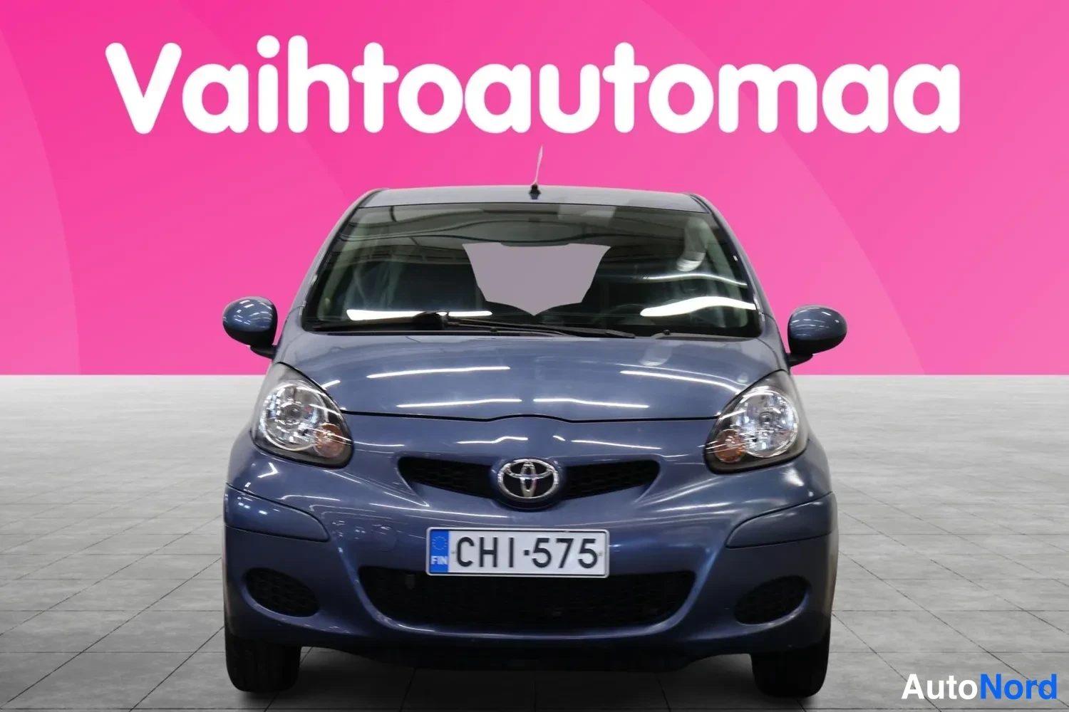Edestä: Sininen Toyota Aygo suoraan edestä, kaksiosaiset CD-keilat ja rekisterikilpi näkyvät; pinkki tausta, valkoinen Vaihtoautomaan logo ylhäällä.