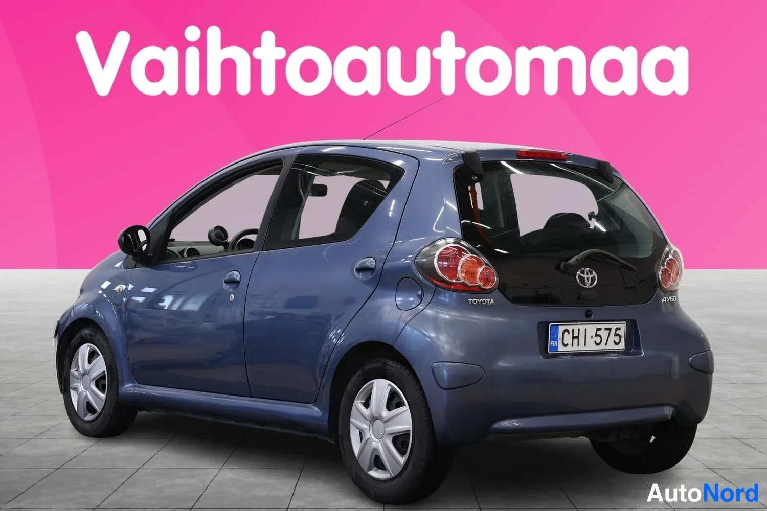 Takaviisto: Sininen Toyota Aygo takaa vasemmalta kolmannelta kuvattuna, rekisteritunnus, vaaleanharmaa studiovaaka.
