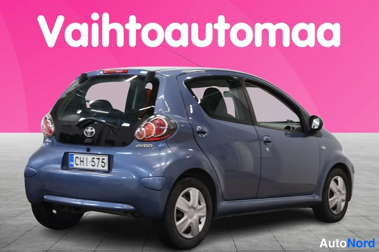 Takaviisto: Sininen Toyota Aygo takaa oikealta kolmannelta kuvattuna, rekisteritunnus, vaaleanharmaa studiovaaka.