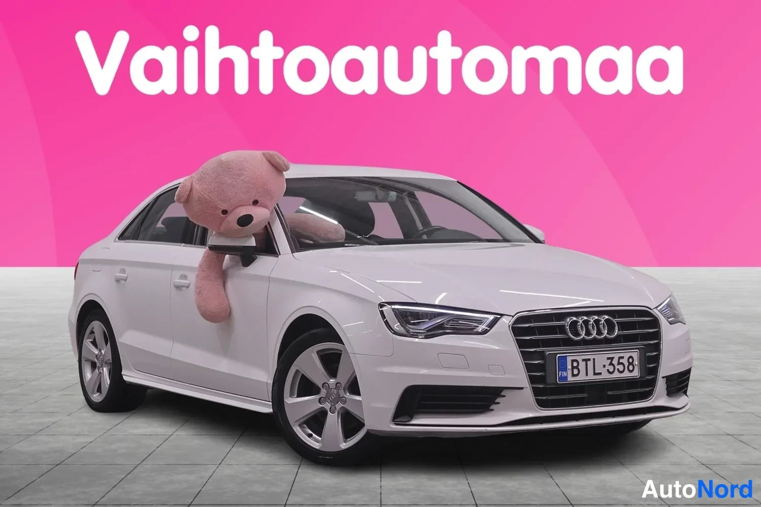 Etuviisto, nokka oikealle: Valkoinen Audi-sedan kolmineljännäksessä edestä oikealta, rekisterikilpi, pehmolelu takanäytön takana, Vahtoautoma-mainos