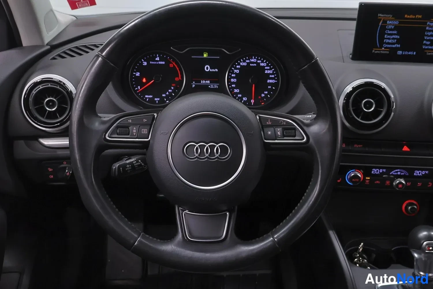 Edestä: Ohjaamoon suunnattu näkymä: Audi-ohjauspyörä, mittaristossa 0 km/h ja P-vaihde, keskikonsolissa näyttö ja automaattivaihteiston valitsin.