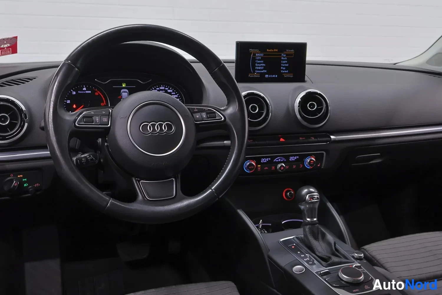 Edestä: Ohjaamo kuljettajan puolelta: Audi-ohjauspyörä, nostettu keskellä oleva infotainment-näyttö, kaksivyöhykeilmastointi ja automaattivaihteiston valitsin.