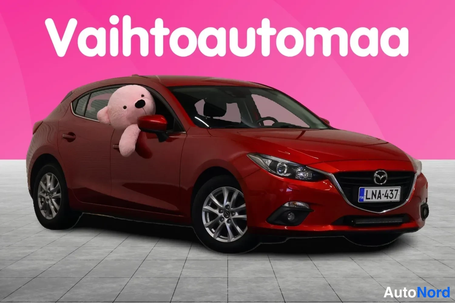 Etuviisto, nokka oikealle: Punainen Mazda 3 -henkilöauto edestä oikealta vasemmalle kuvattuna, rekisterikilpi, vaaleanpunainen pehmolelu takapyyhkimossa ikkunassa, taustalla vaalea
