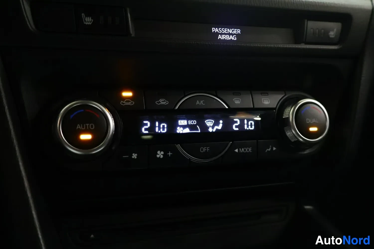 Muu: Autojen ilmastointipaneeli, jossa lukee 21 astetta molemmissa säätimissä, AUTO ja DUAL -merkit sekä A/C-painike ja teksti PASSENGER AIRBAG.
