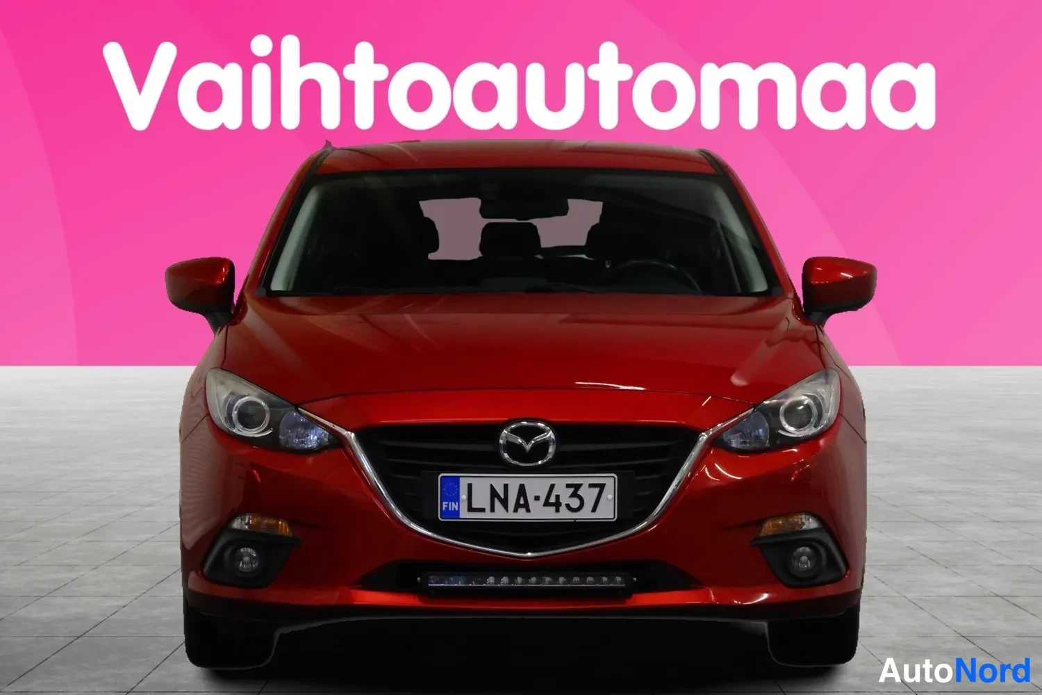 Edestä: Punainen Mazda 3 -henkilöauto suoraan edestä kuvattuna, etusäleikössä Mazda-merkki, rekisterikilpi, taustalla vaaleanpunainen mainoskankaan teksti Vaihtoautomaa