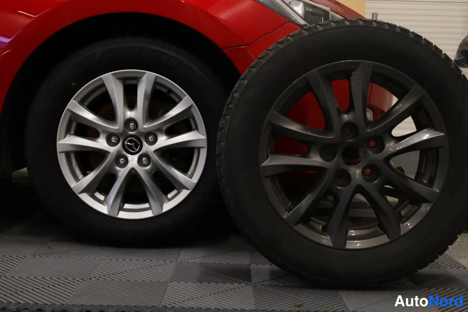 Sivulta: Kaksi pyörää seisomassa lattialla punaisen auton vieressä. Vasen pyörä on sileällä renkaalla ja hopeanharmaalla vanteella (Mazda-logo). Oikealla pyörässä talvir