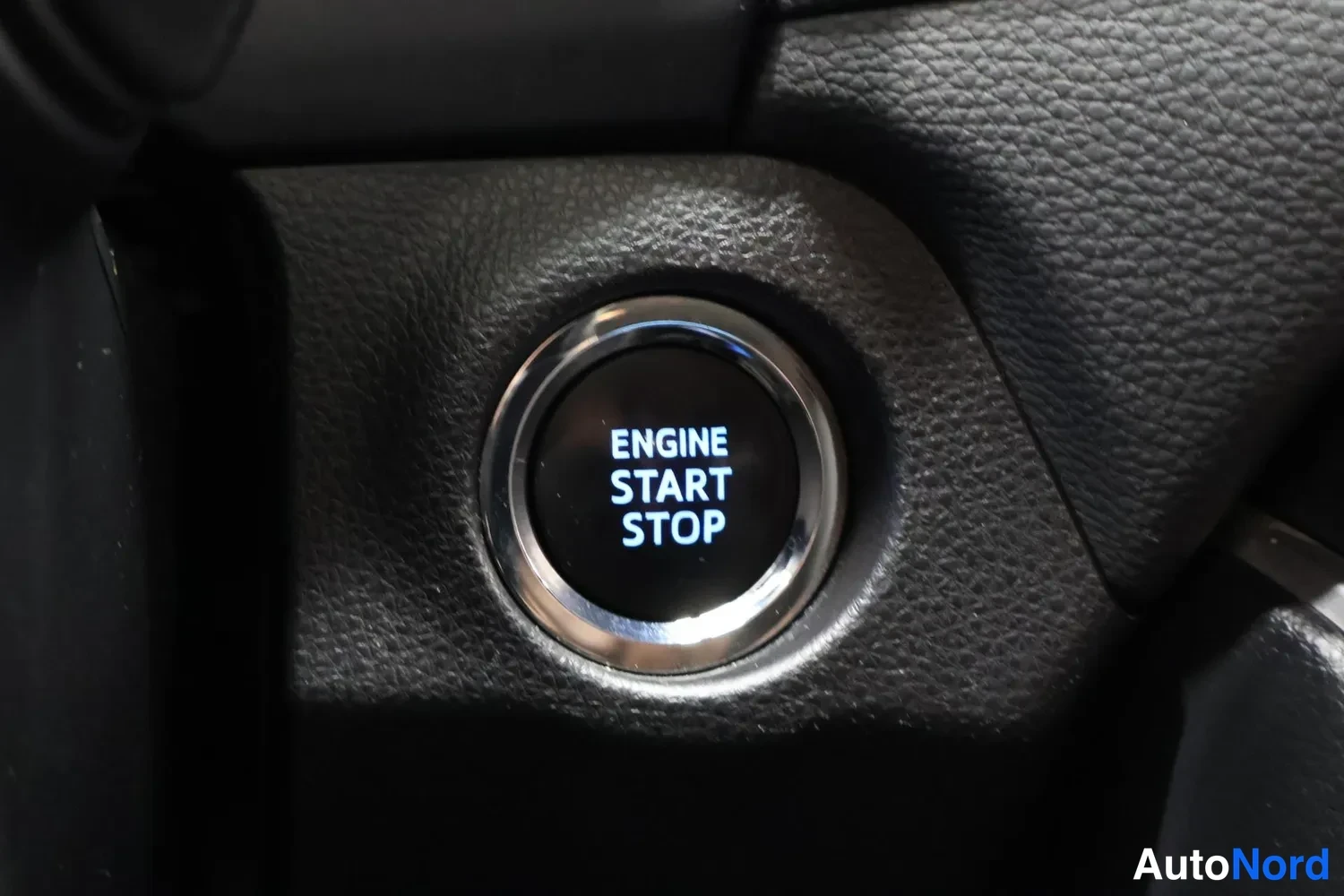 Yksityiskohta: Painike pyörivällä hopeareunuksella, jossa lukee ENGINE START STOP, mustan muovipinnan keskellä.
