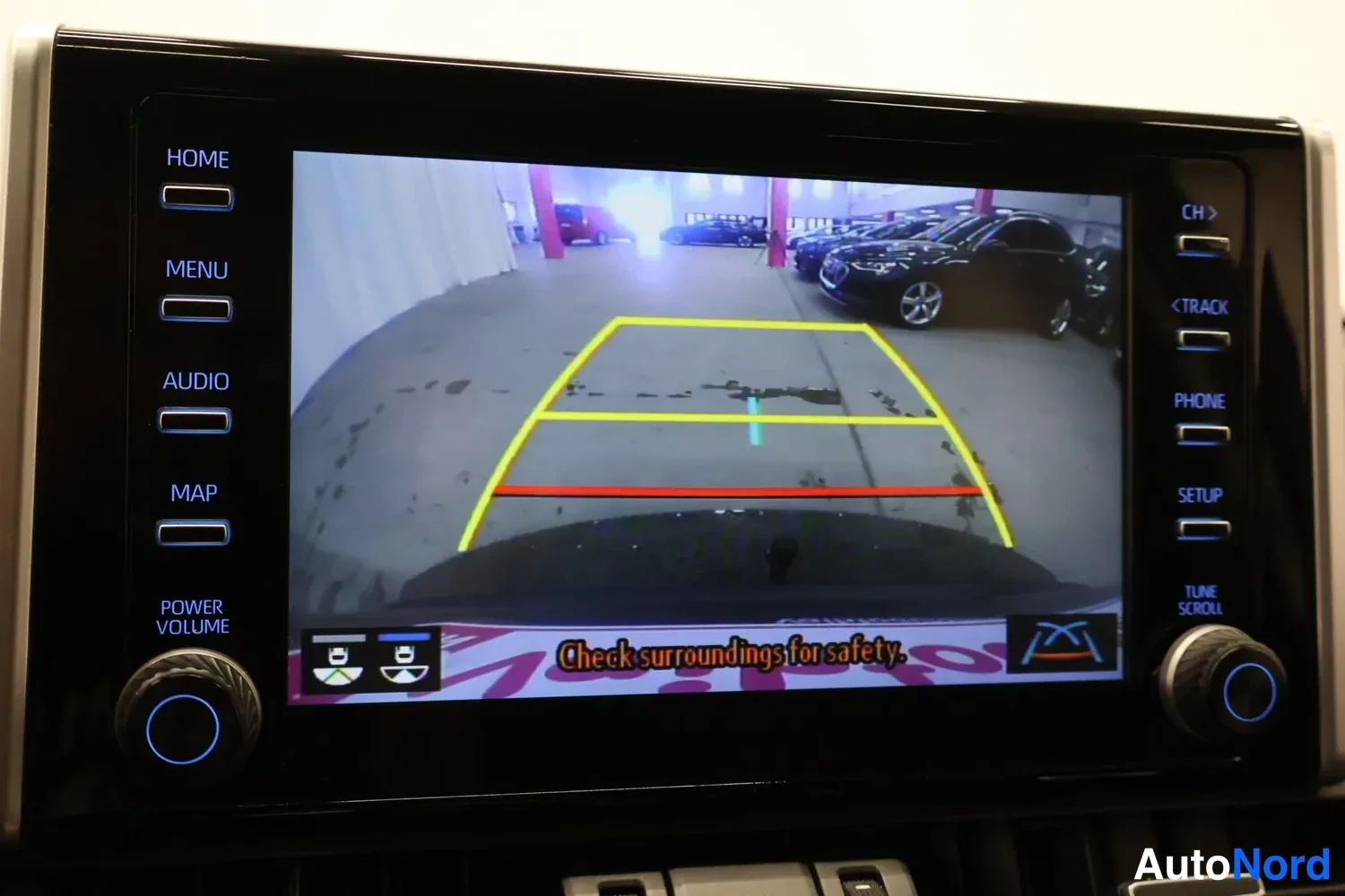 Yksityiskohta: Auton näyttö, jossa näkyy peruutuskameran kuva parkkihallista ja keltaisen-punaisen ohjauslinjat sekä teksti 'Check surroundings for safety.'