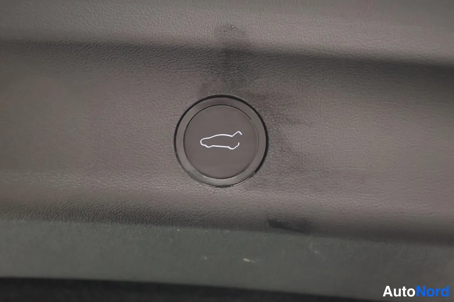 Yksityiskohta: Autoa esittävä valaistu painike tummanharmaassa panelissa, tarkoitus todennäköisesti käynnistää jokin autoon liittyvä toiminto.