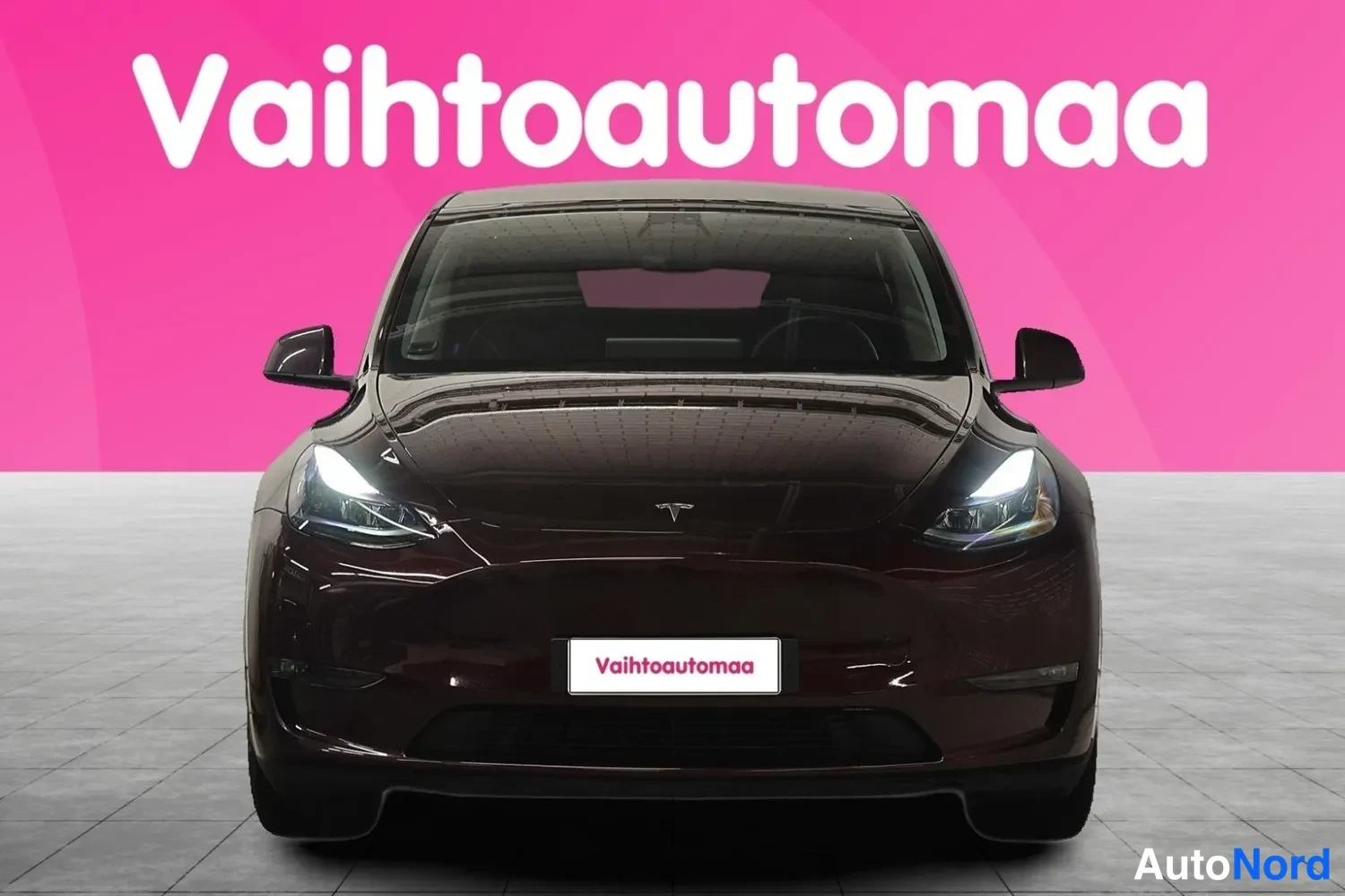 Edestä: Tummanpunainen Tesla-merkkinen sähköauto suoraan edestä kuvattuna, valot päällä, etuvempaimessa Vaihtoautoma-kyltti, taustana pinkki kulissi.