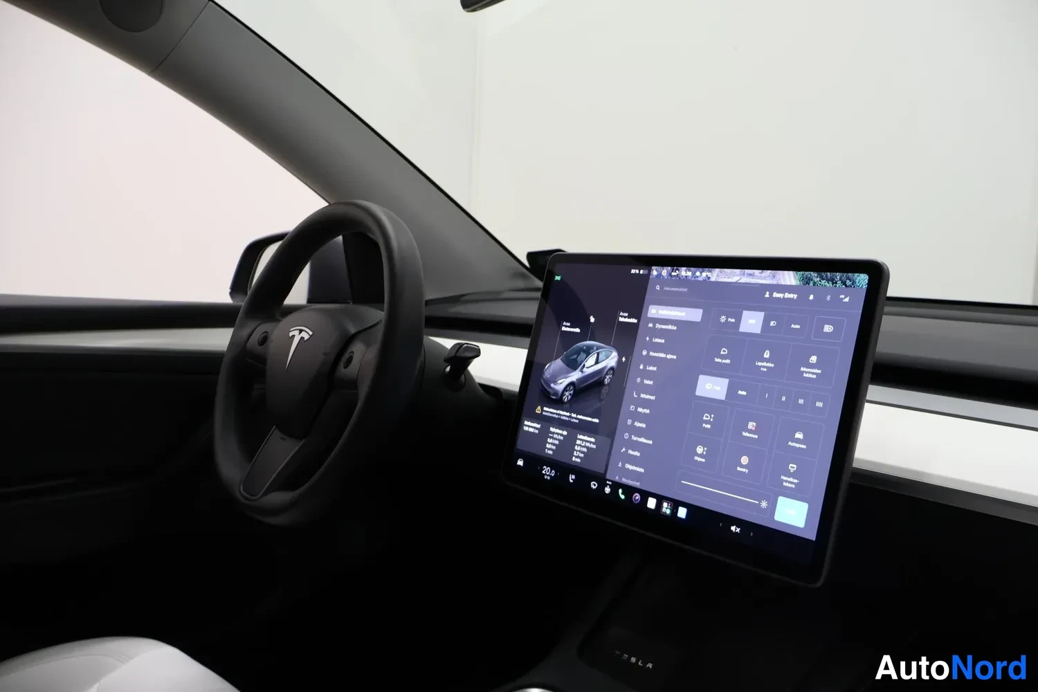 Muu: Teslaan kuljettajan puolelta viistosti kuvattu sisätilanne: ohjauspyörä Tesla-logolla vasemmalla, iso kosketusnäyttö näkyvillä valikkoineen ja kojelaudan valkoi