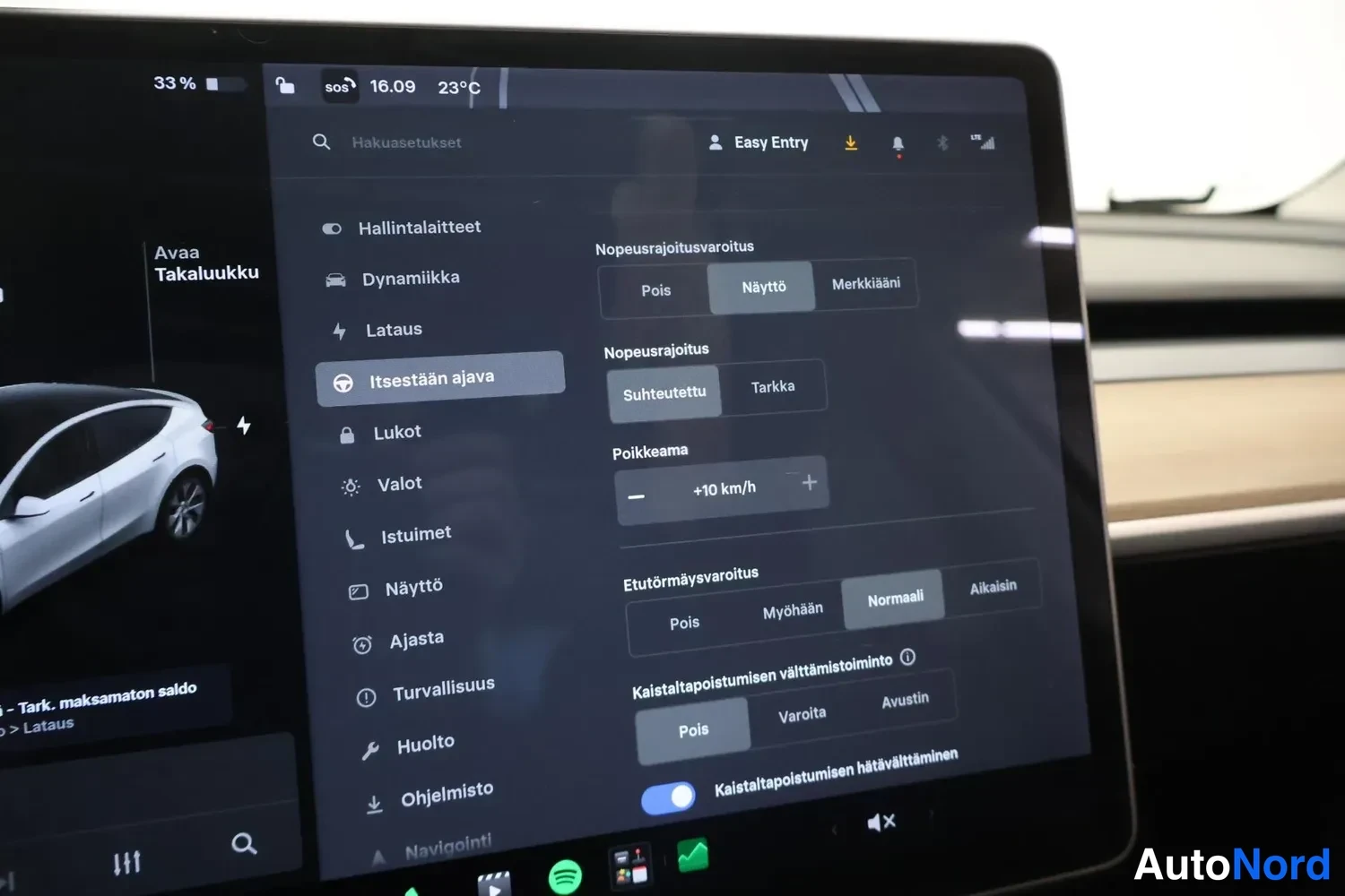 Yksityiskohta: Tesla-ajoneuvon kosketusnäyttö, jossa näkyy Autopilot-asetukset: nopeusrajoitus asetettu tilaan Näyttö, arvo suhteutettu ja poikkeama +10 km/h, sekä etutörmäysv