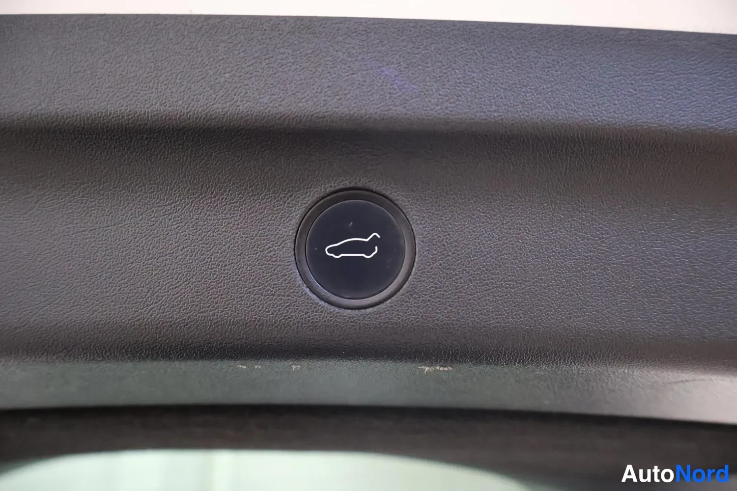Yksityiskohta: A close-up of a black circular button featuring a white tailgate/trunk icon.