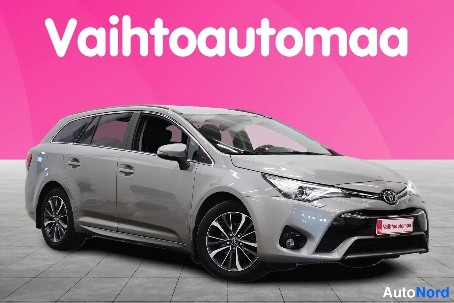 Etuviisto, nokka oikealle: Hopeanharmaa Toyota Avensis farmari etuviistöstä oikealta, valokuvattu studiossa vaaleanharmaalla lattialla ja pinkillä taustalla.