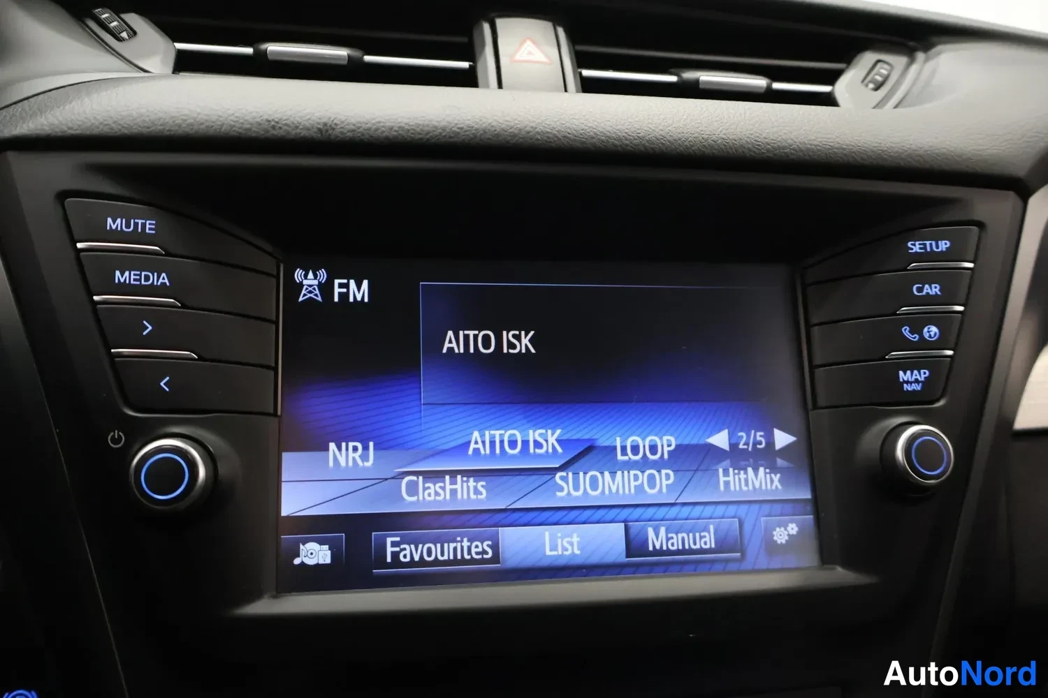 Yksityiskohta: Kojelaudan infotainment-näytön kuva: radiossa on FM-viritetty kanava AITO ISK, valikoimissa NRJ, ClasHits, LOOP, SUOMIPOP ja HitMix, alavalikot Favourites, List