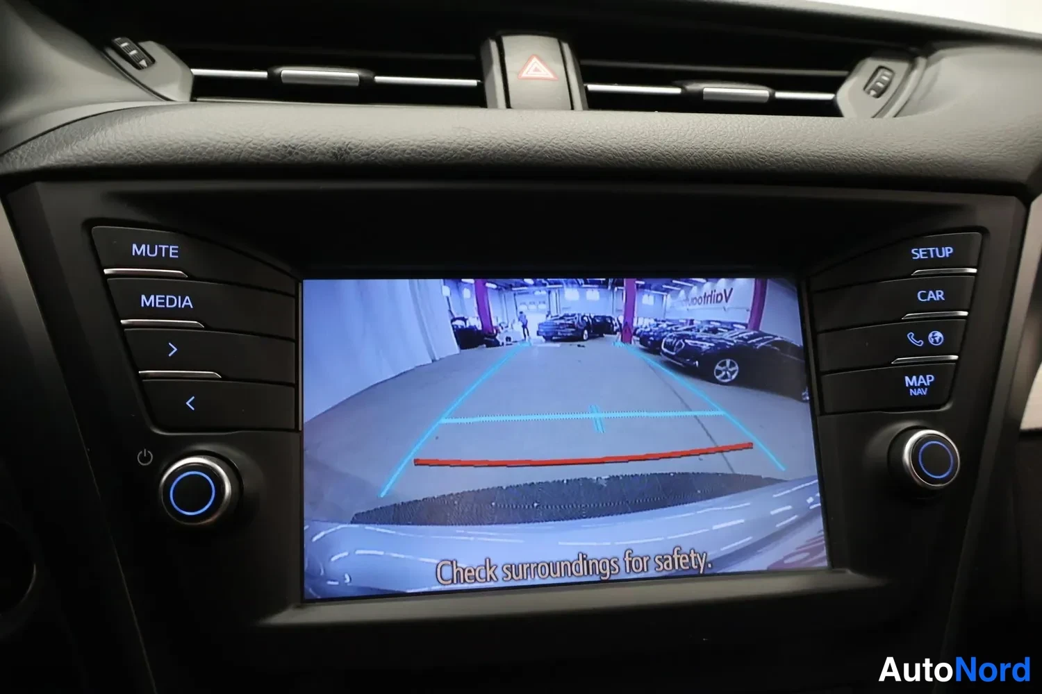 Yksityiskohta: Auton medianäyttö näyttää peruutuskameran kuvaa hallitilasta, apuviivoineen ja tekstillä Check surroundings for safety.