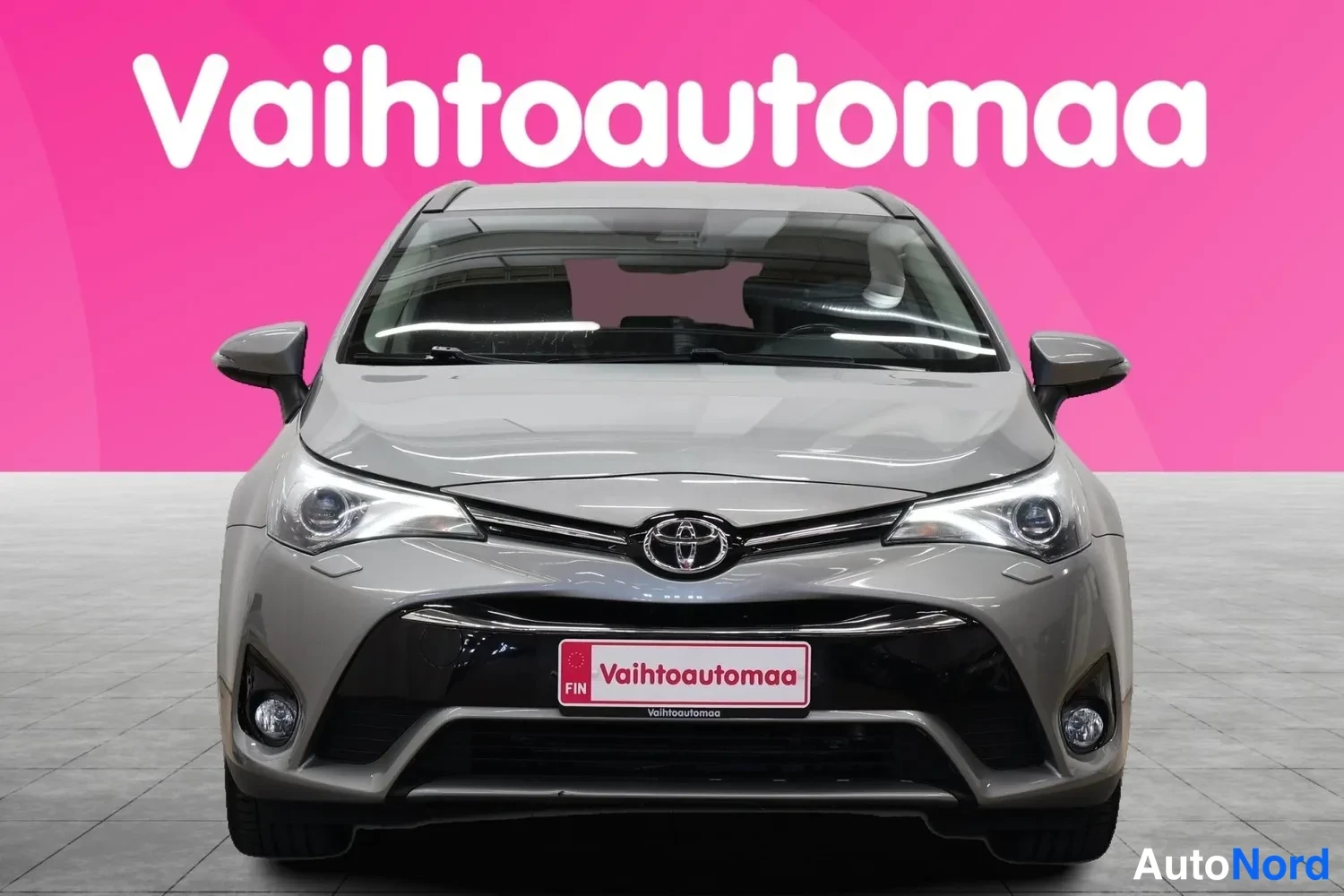 Edestä: Toyota Avensis farmarin keula suoraan edestä: etupuskuri, Mazda-merkki, ajovalot ja sumuvalot näkyvät, taustalla pinkki seinä ja vaaleanharmaa lattia.