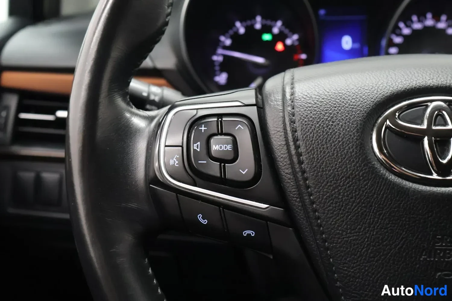 Muu: Ratin vasen puoli lähikuva: Toyota-merkki ja ääniohjainnäppäimet ('MODE' ja puhelinnäppäimet), taustalla mittaristo.